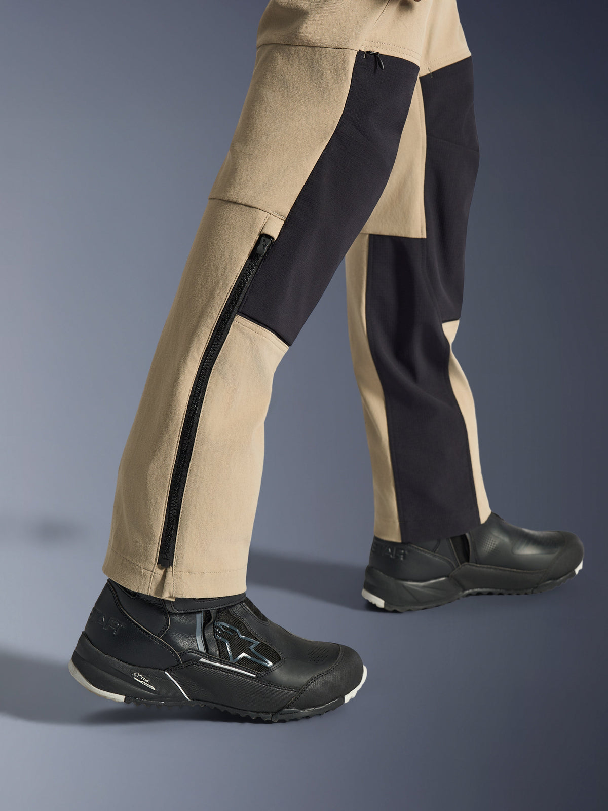 Alpinestars Woman Stella Flex-AST Explorer Pants, motocyklowe spodnie adventure, piaskowe i czarne, zbliżenie na dolną część nogawek i kostki z długim bocznym zamkiem błyskawicznym i czarnymi panelami wzmacniającymi, noszone z czarnymi butami motocyklowymi Alpinestars