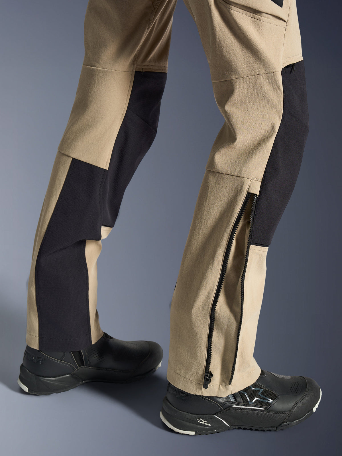 Alpinestars Stella Flex-AST Explorer Pants, damskie spodnie motocyklowe, piaskowe i czarne, zbliżenie na dolną część nogawek pokazujące dół z zamkiem błyskawicznym ułatwiającym dopasowanie butów, noszone z czarnymi butami motocyklowymi Alpinestars, z wytrzymałymi panelami z materiału ripstop i oddychającej, elastycznej tkaniny
