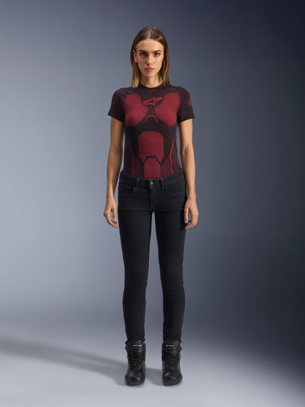 Alpinestars Daisy V3 Women's Riding Denim, Rinse Black, dopasowane spodnie motocyklowe slim-fit, prezentowane przez modelkę w technicznej koszulce z czerwono-czarnym wzorem i czarnych butach motocyklowych, codzienna miejska odzież motocyklowa o kobiecej sylwetce i fasonie z wysokim stanem