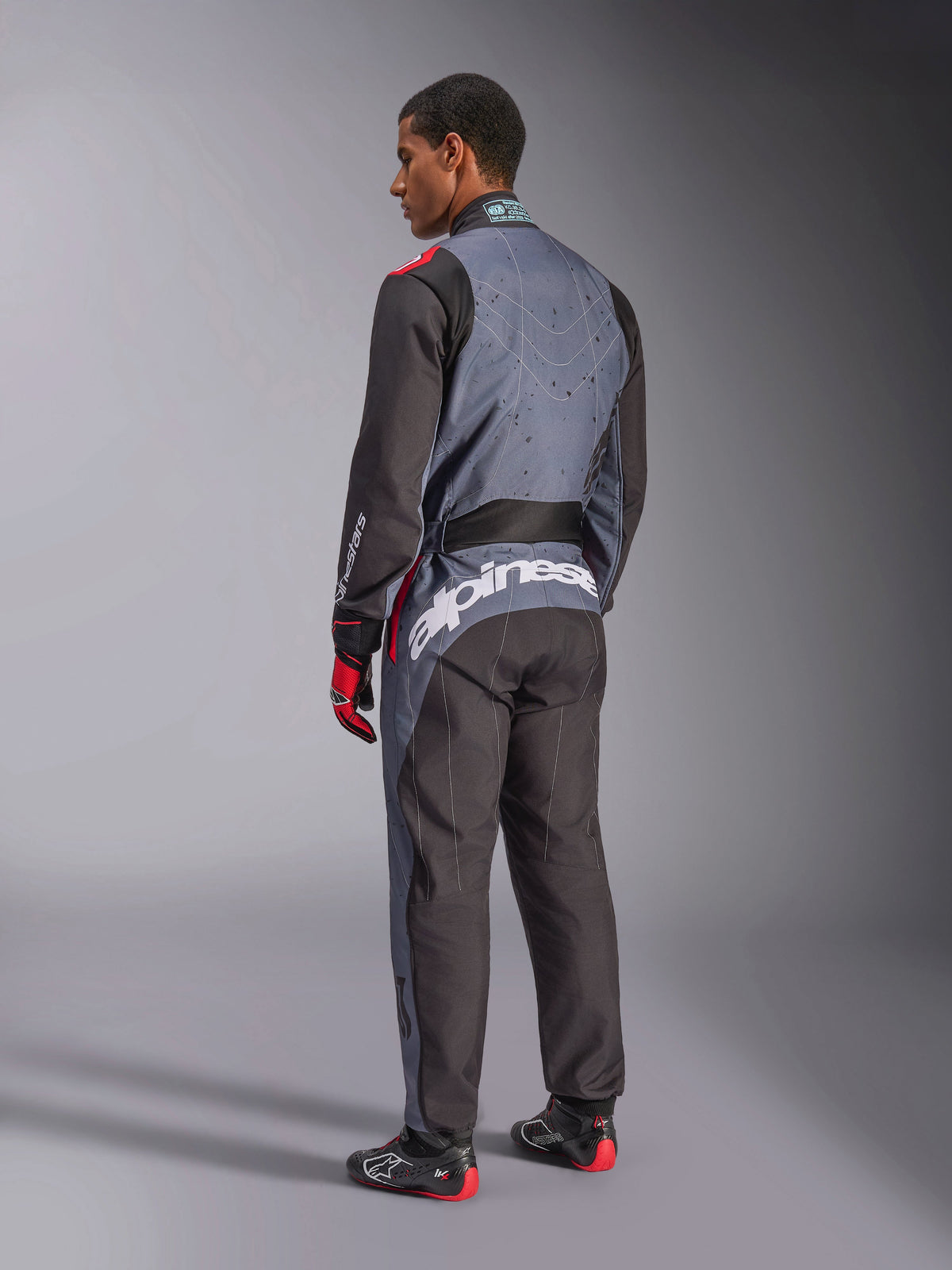 Widok z tyłu modelu w kombinezonie Alpinestars KMX-9 V3 Graphic 6, kombinezon kartingowy, kolor antracytowo-czarno-czerwony, z wyraźnym białym brandingiem Alpinestars na dolnej części pleców, czarno-szary panelowy design z czerwonymi akcentami, prezentowany z pasującymi rękawicami i butami wyścigowymi.