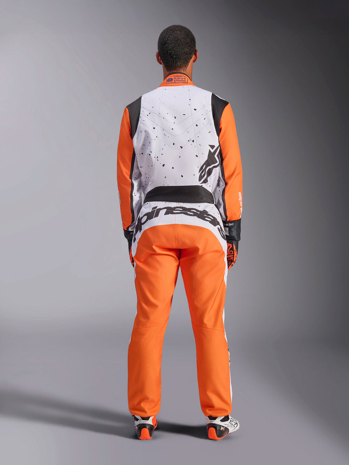 Widok z tyłu zawodnika w kombinezonie Alpinestars KMX-9 V3 Graphic 6 w kolorze biało-pomarańczowo-czarnym, biała górna część pleców z czarną nakrapianą grafiką, pomarańczowe rękawy i spodnie, duże czarne logo Alpinestars na dole pleców, w zestawieniu z rękawicami i butami wyścigowymi do kartingu.