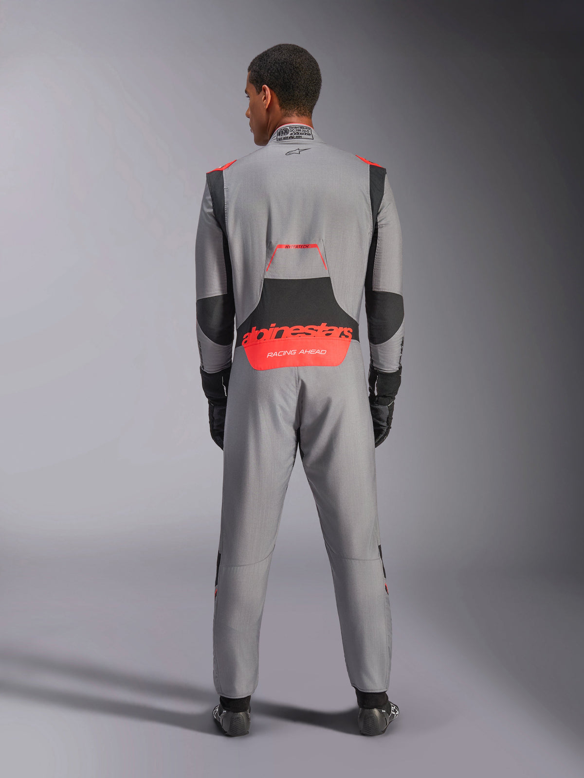 Kombinezon Alpinestars Hypertech V3, profesjonalny kombinezon samochodowy, szary z czarnymi i czerwonymi akcentami Fluo, widok z tyłu na modelu z czerwonym brandingiem Alpinestars w dolnej części pleców, lekka konstrukcja z Nomexu dla wydajności na torze.