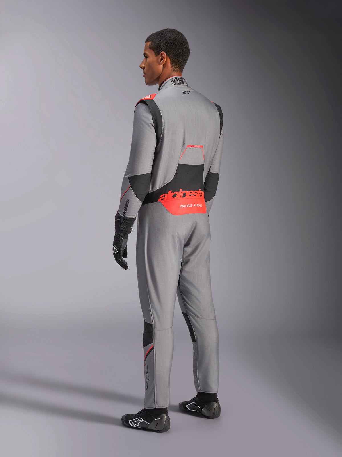 Kombinezon Alpinestars Hypertech V3 w kolorze Mid Gray Black Red Fluo, widok z tyłu pod kątem trzech czwartych, z wyraźnym czerwonym brandingiem Alpinestars w dolnej części pleców, czarnymi elastycznymi panelami i dopasowanym sportowym krojem do profesjonalnych wyścigów samochodowych.