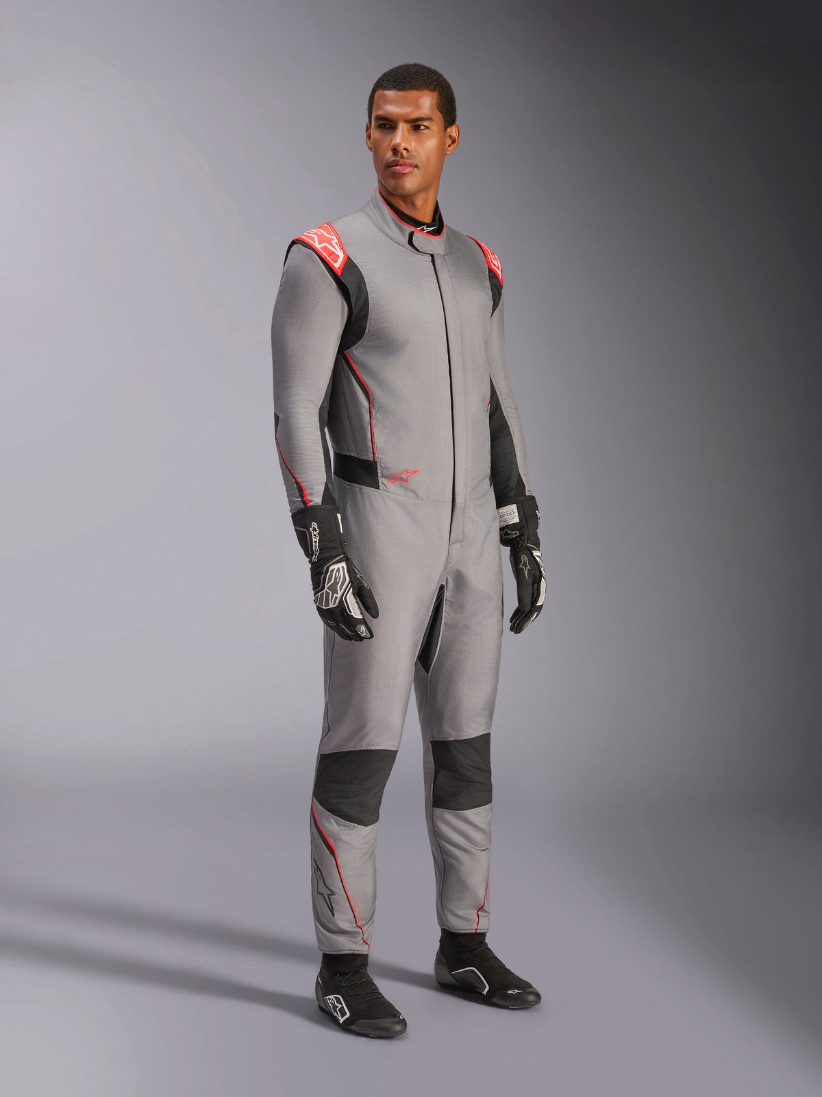 Męski model w kombinezonie Alpinestars Hypertech V3 w kolorze Mid Gray z czarnymi i czerwonymi akcentami Fluo, profesjonalny samochodowy kombinezon wyścigowy zestawiony z czarnymi rękawicami i butami wyścigowymi, charakteryzujący się lekką dwuwarstwową konstrukcją Nomex z czerwonymi detalami na ramionach i sportowym krojem