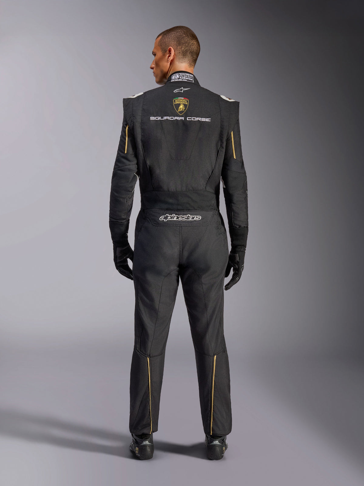 Alpinestars Limited Edition Lamborghini GP Tech V4 Suit FIA, Auto Racing Suit, Black with Gold and Silver accents, rear view showing Lamborghini Squadra Corse branding, Alpinestars logo, and gold piping on sleeves and legs, professional racing gear
