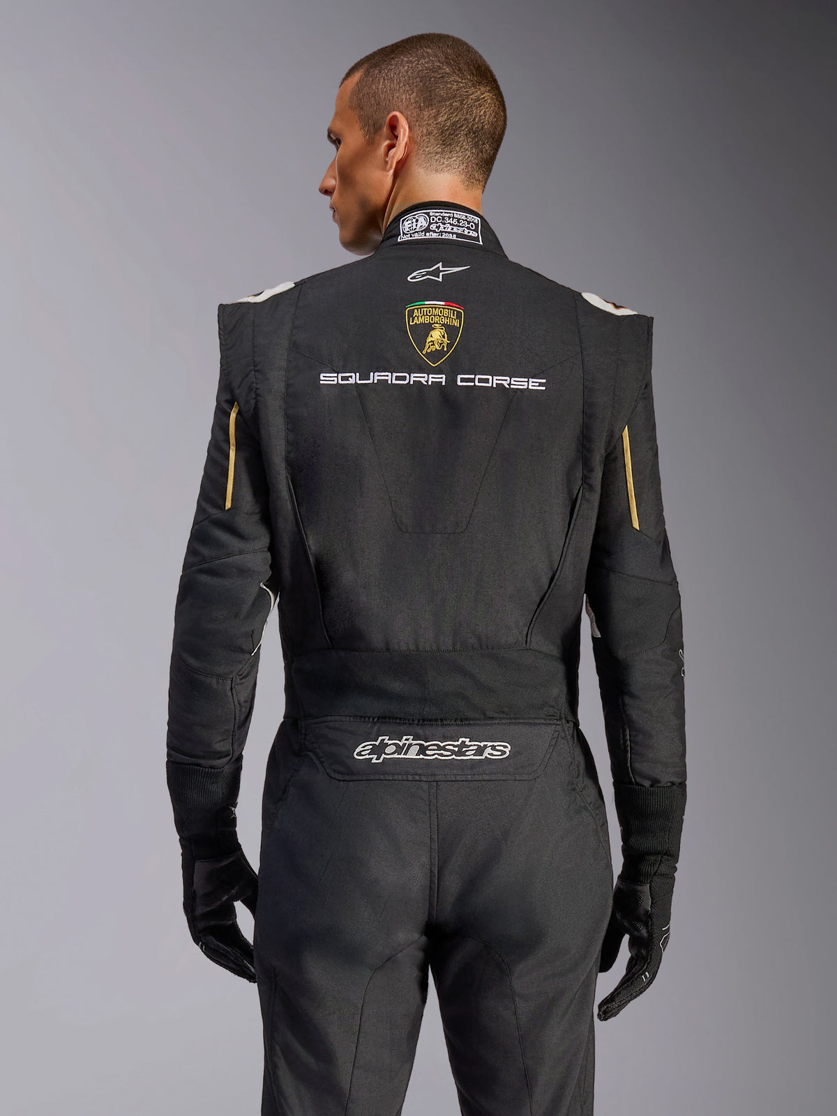 Rear view of the Alpinestars Limited Edition Lamborghini GP Tech V4 Suit, Black with Gold accents, featuring Automobili Lamborghini shield and 'SQUADRA CORSE' text on the back, professional FIA-certified racing suit for automotive competition