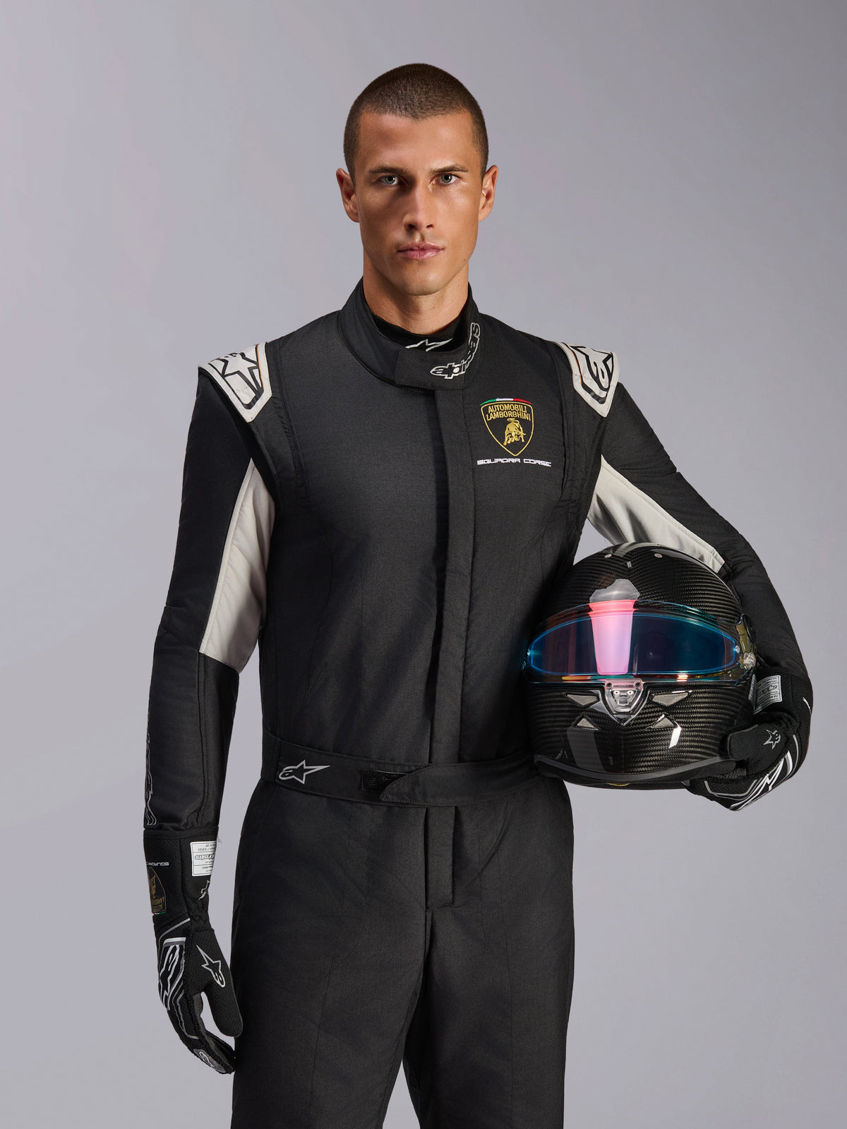 Alpinestars Limited Edition Lamborghini GP Tech V4 Suit, Auto Racing Suit, Black Silver and Gold, worn by a model holding a carbon fiber helmet with an iridescent visor, featuring Lamborghini Squadra Corse branding and white shoulder accents