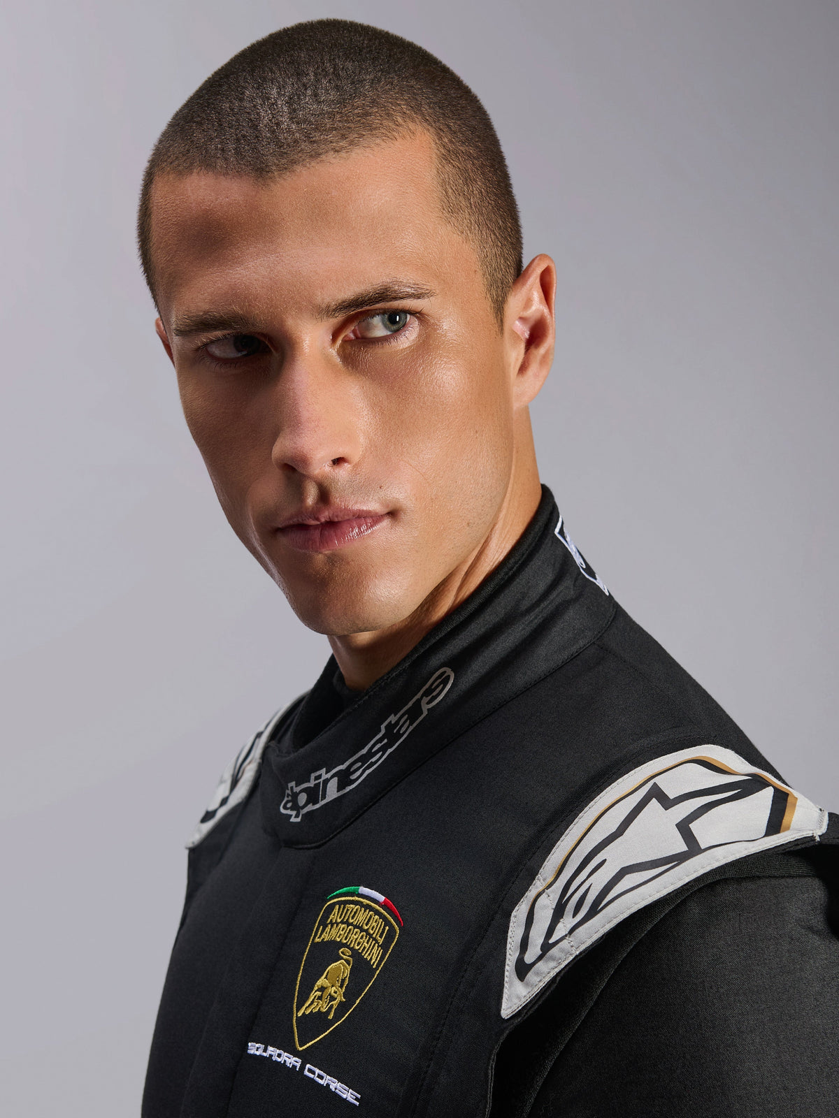 Close-up of a male model wearing the Limited Edition Lamborghini GP Tech V4 Suit, Black Silver and Gold, featuring Automobili Lamborghini Squadra Corse and Alpinestars logos, professional FIA-certified auto racing gear