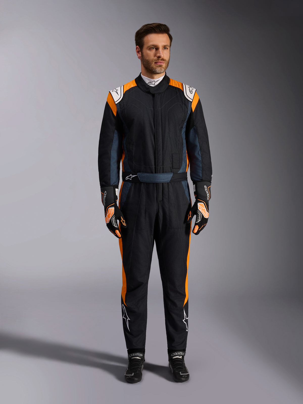 Alpinestars GP Pro Comp V2 FIA Suit, Black Asphalt and Orange Fluo auto racing suit, male model wearing full protective gear with matching racing gloves and shoes, features white Alpinestars logos and vibrant orange side panels, professional motorsport apparel