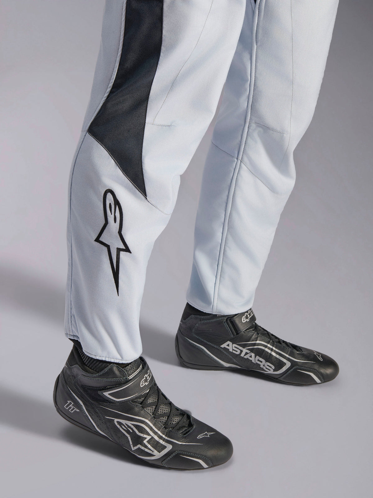 Alpinestars GP Pro Comp V2 FIA, kombinezon wyścigowy, szary i czarny, logo gwiazdy na nogawce i czarne buty wyścigowe.