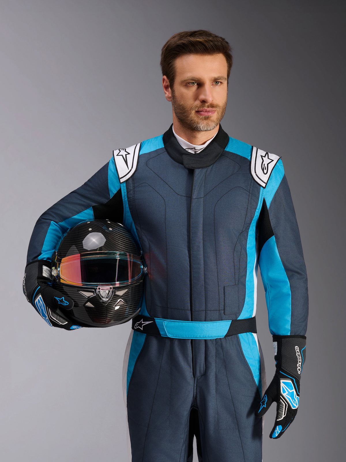 Alpinestars GP Pro Comp V2 Suit - FIA, Auto Racing Suit in Asphalt Cyan and White, worn by a man holding a helmet.