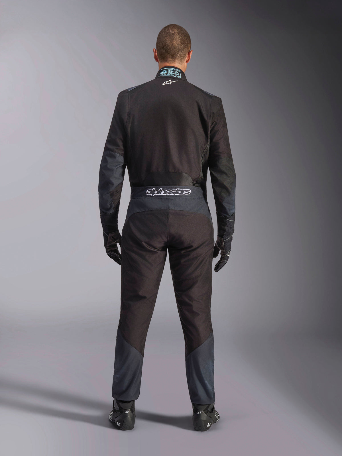 Kombinezon Alpinestars KMX-5 V3, kombinezon kartingowy, czarny i ciemnoszary (Dark Shadow), widok z tyłu na kierowcę w pełnym kombinezonie z pasującymi rękawicami i butami, logo Alpinestars na pasku, ergonomiczne panele
