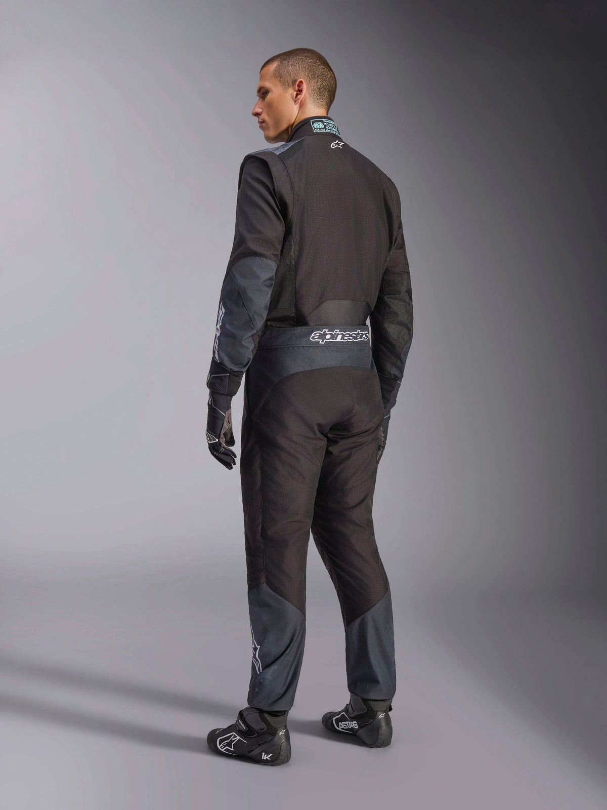 Kombinezon Alpinestars KMX-5 V3 w kolorze Black Dark Shadow, widok z tyłu na modela w pełnym stroju kartingowym z dopasowanymi rękawicami i butami wyścigowymi, logo Alpinestars na pasie, ciemnoszare akcenty, profesjonalny sprzęt kartingowy.