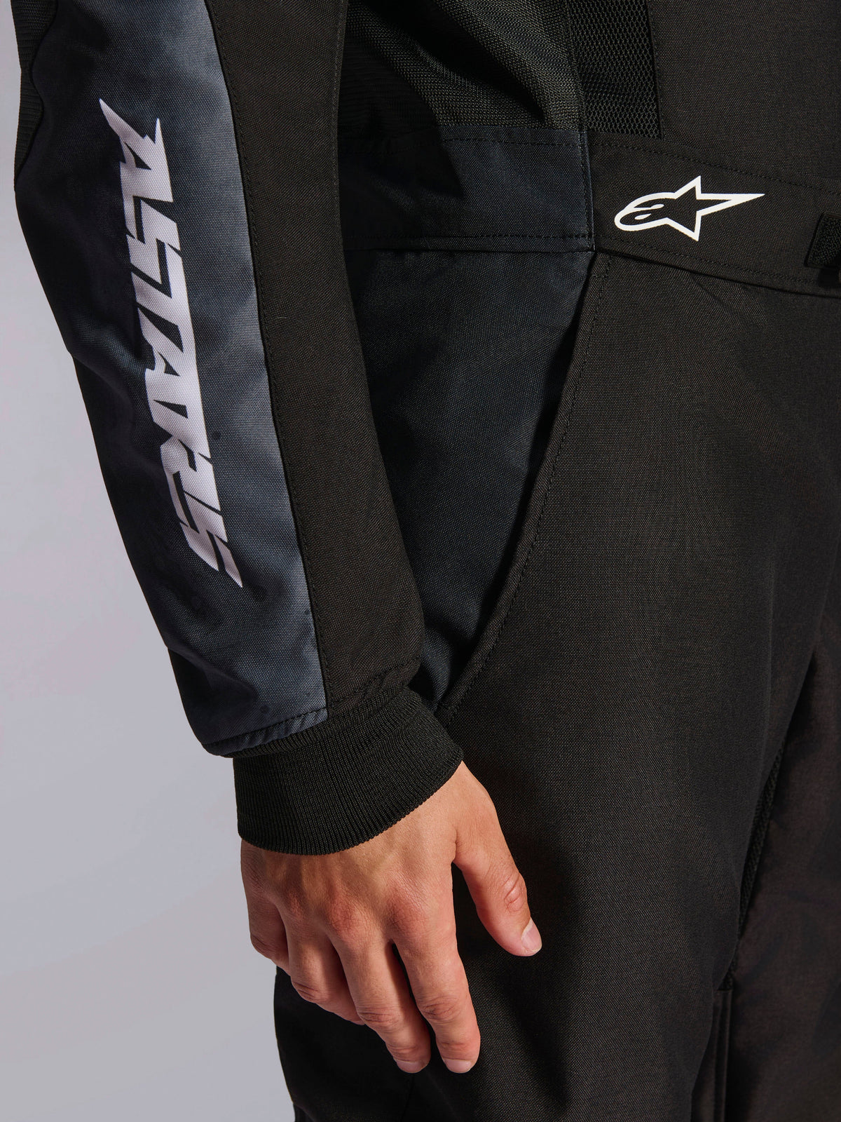 Kombinezon Alpinestars KMX-5 V3, kombinezon kartingowy, czarny i Dark Shadow, zbliżenie na biodro i ramię, biały napis ASTARS na rękawie, białe logo w pasie, z panelami z siatki dla wentylacji i dzianinowym mankietem