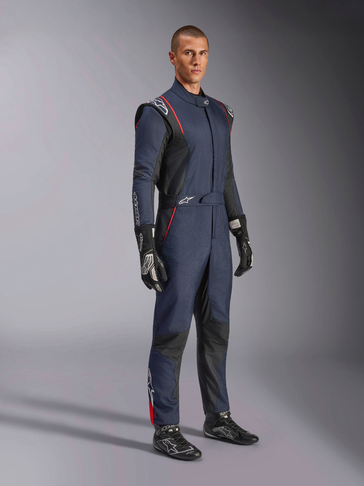 Kombinezon Alpinestars GP Tech V4, jednoczęściowy samochodowy kombinezon wyścigowy, granatowy z czarnymi i czerwonymi elementami, prezentowany przez modela w czarnych rękawicach i butach wyścigowych, czarne panele i czerwone lamówki, profesjonalna odzież motorsportowa