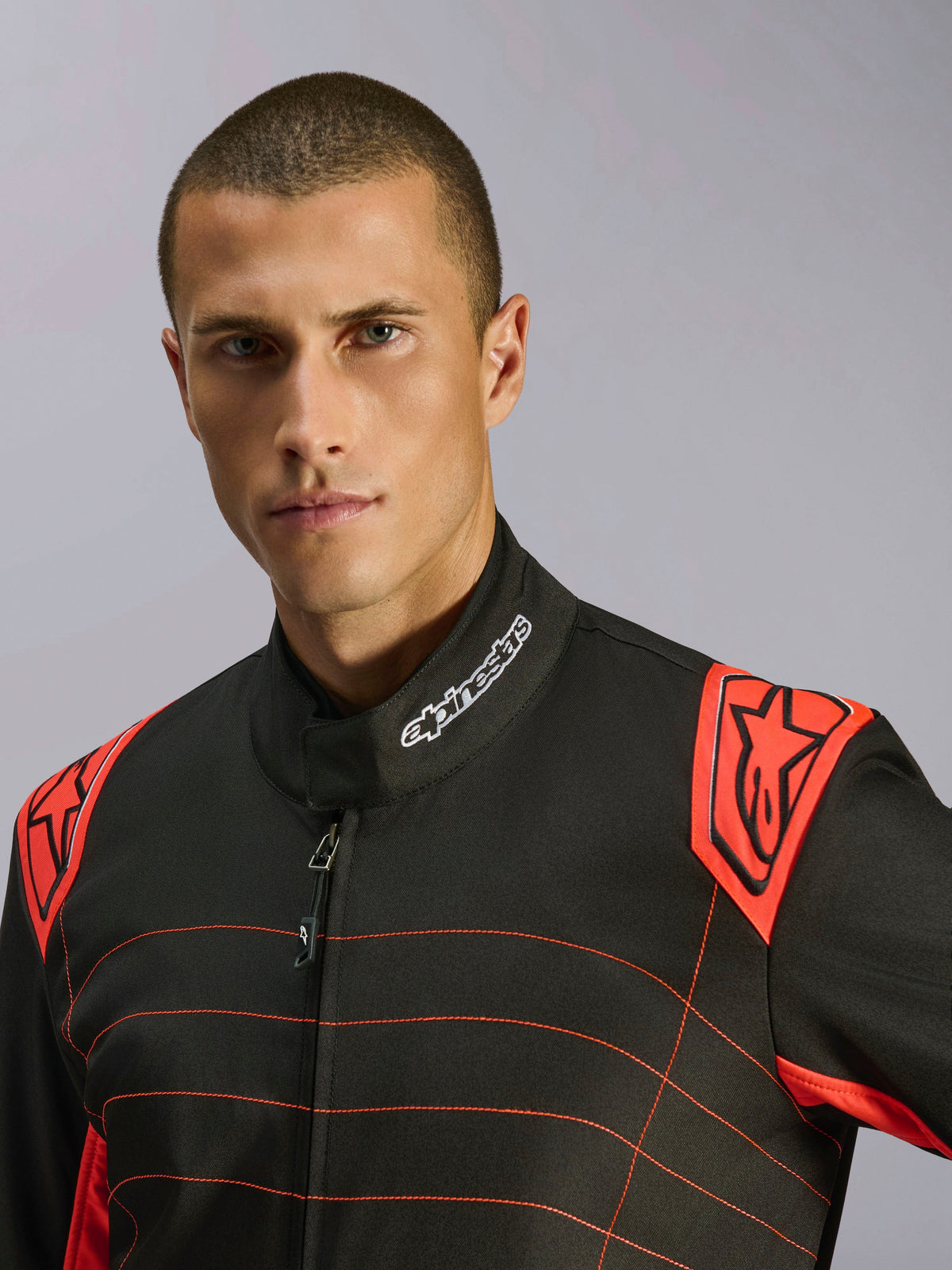 Zbliżenie na kierowcę w kombinezonie Alpinestars KMX-9 V3 w kolorystyce Black Red Fluo, z haftowanym logo na kołnierzu i czerwonymi naramiennikami z brandingiem Alpinestars, techniczny design odzieży kartingowej
