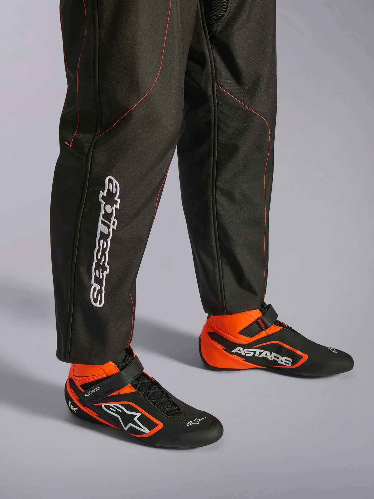 Kombinezon Alpinestars KMX-9 V3, kombinezon kartingowy, czarny i czerwony fluo, szczegółowy widok nogawek i butów wyścigowych, czarny materiał z czerwonymi przeszyciami fluo, pionowe logo Alpinestars na łydce, czarno-pomarańczowe buty wyścigowe, profesjonalny karting