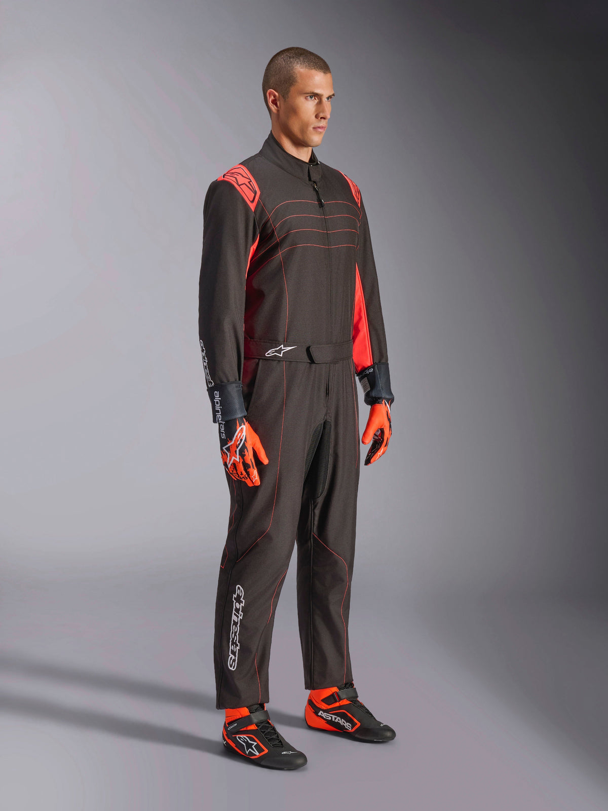 Alpinestars KMX-9 V3 Suit, kombinezon kartingowy, czarno-czerwony fluorescencyjny, prezentowany przez modela z pomarańczowymi rękawicami i czarnymi butami wyścigowymi, czerwone przeszycia, akcenty na ramionach i białe logo, sportowy design do profesjonalnych wyścigów