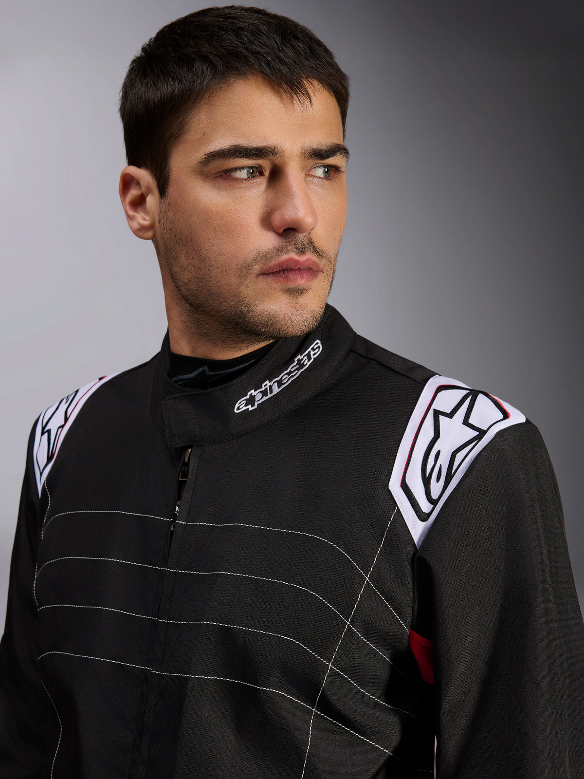 Alpinestars KMX-9 V3 Suit, czarny kombinezon kartingowy prezentowany przez modela, zbliżenie na klatkę piersiową i ramiona, białe logo na kołnierzu i naramiennikach, białe ozdobne przeszycia, profesjonalny design motorsportowy