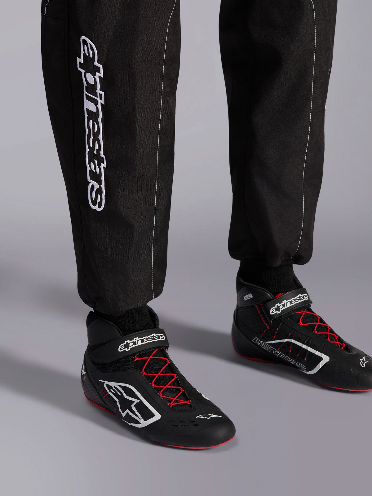 Kombinezon Alpinestars KMX-9 V3, kombinezon kartingowy, czarny, czerwony i biały, widok szczegółowy na dolną część nogawek i buty kartingowe, białe logo Alpinestars z boku, czarne buty z czerwonymi sznurowadłami i białymi akcentami, profesjonalny sprzęt wyścigowy