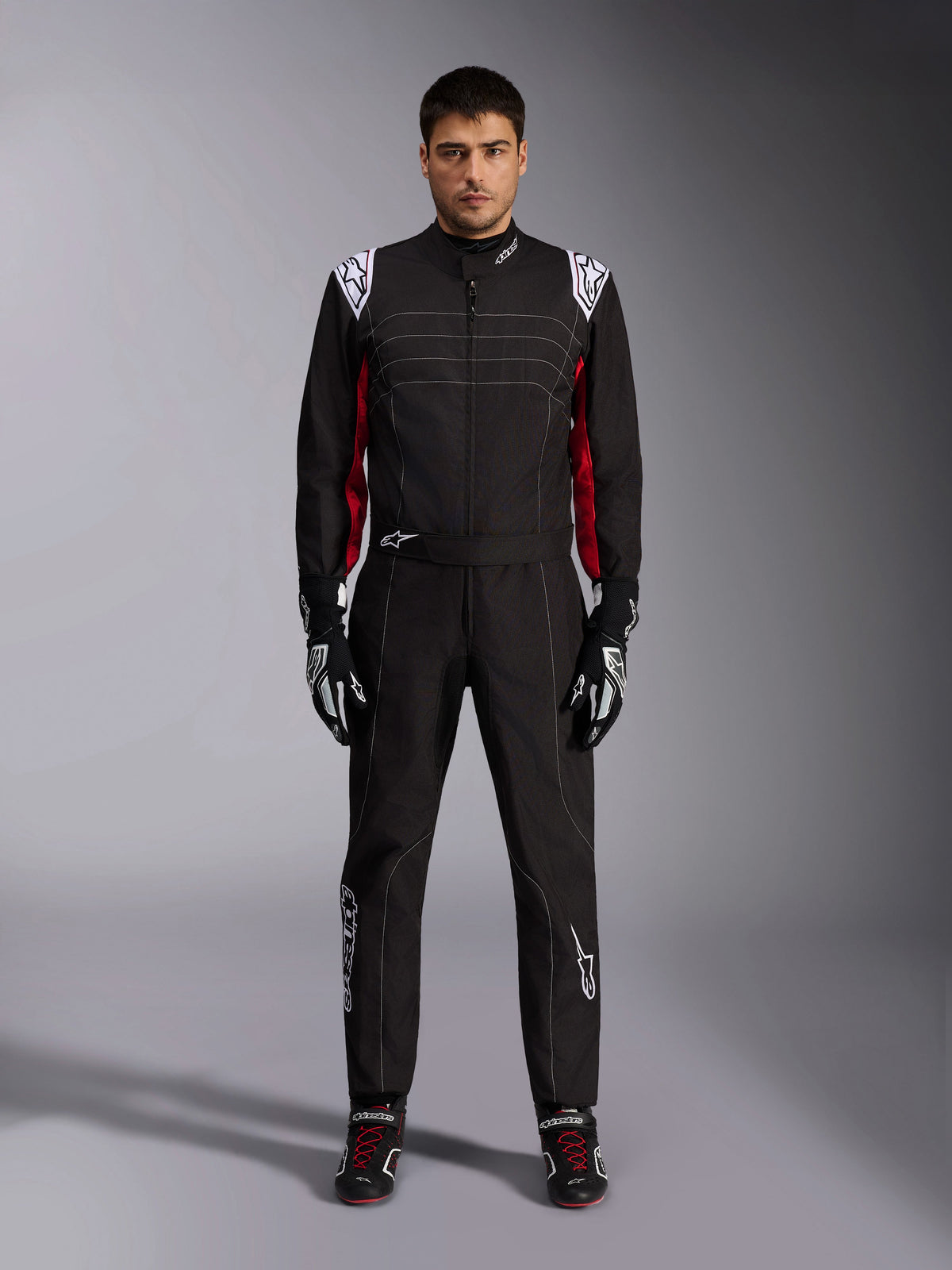 Kombinezon kartingowy Alpinestars KMX-9 V3 w kolorze czarno-czerwono-białym, widok z przodu na całą sylwetkę modela ubranego w jednoczęściowy kombinezon z dopasowanymi rękawicami i butami wyścigowymi, białe logo Alpinestars na ramionach i czerwone akcenty
