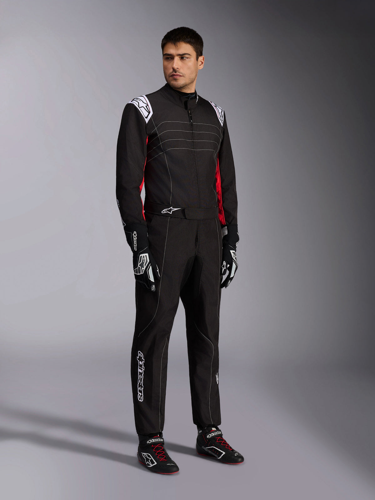 Kombinezon Alpinestars KMX-9 V3, czarno-czerwono-biały kombinezon kartingowy prezentowany przez modela, białe naramienniki i logo, czerwone akcenty po bokach, dopasowane rękawice i buty wyścigowe, profesjonalna odzież do sportów motorowych