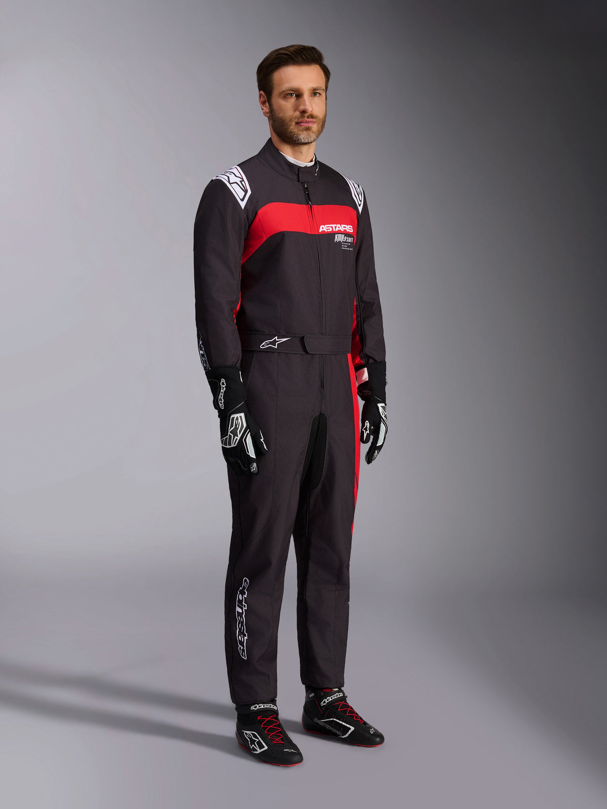 Męski model w kombinezonie Alpinestars KMX-9 V3 Graphic 3, kombinezon kartingowy, czarno-czerwony, z białymi epoletami na ramionach i czerwonym logo na piersi, czarne rękawice wyścigowe i buty z czerwonymi sznurowadłami, profesjonalna odzież motorsportowa.