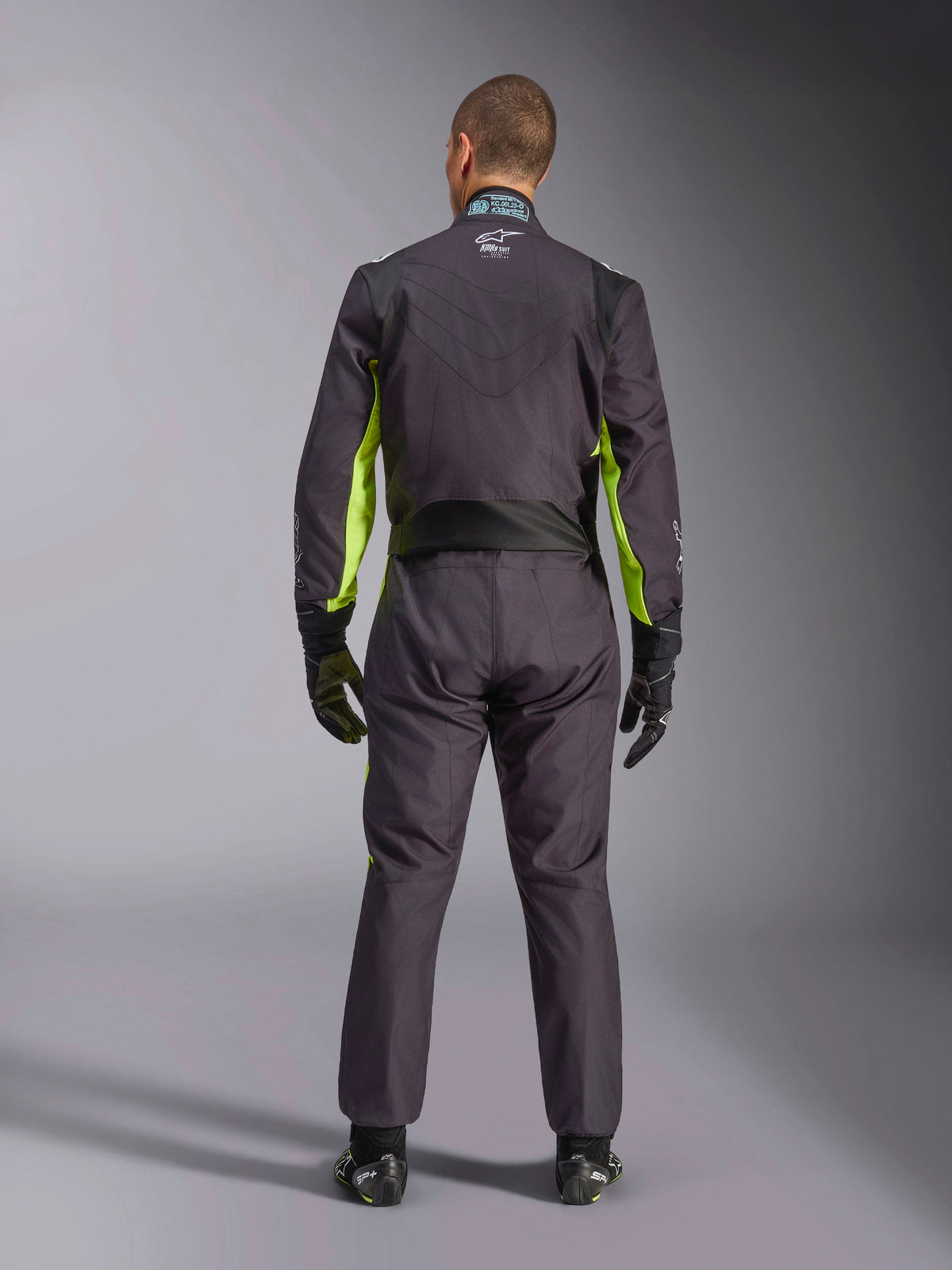 Widok od tyłu na całą sylwetkę osoby ubranej w kombinezon kartingowy Alpinestars KMX-9 V3 Graphic 3 w kolorze czarnym i żółtym fluo, widoczne ergonomiczne panele rozciągliwe na plecach i ramionach, czarne rękawice i buty wyścigowe.
