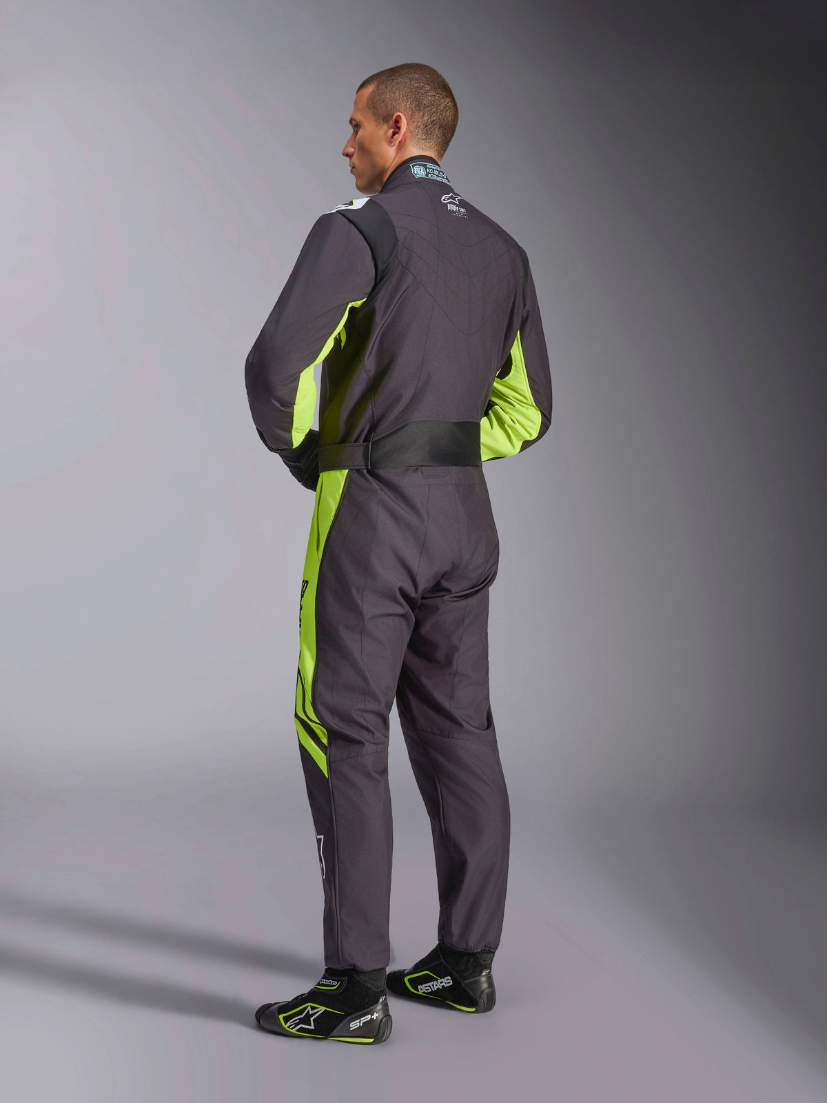 Alpinestars KMX-9 V3 Graphic 3 Suit, kombinezon kartingowy, czarno-żółty fluorescencyjny, widok modelu od tyłu prezentujący jednoczęściową konstrukcję z fluorescencyjnymi żółtymi akcentami po bokach i pod pachami, ergonomiczne elastyczne panele w części lędźwiowej oraz regulowany pas w talii, zestawiony z pasującymi butami wyścigowymi.