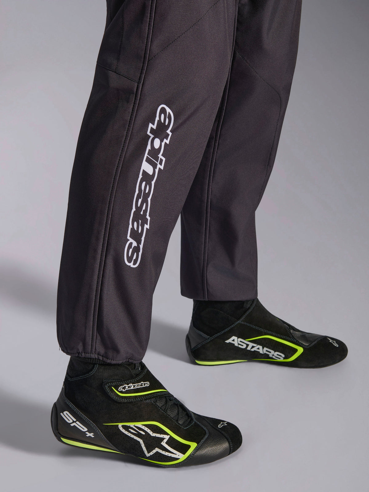 Alpinestars KMX-9 V3 Graphic 3 Suit, kombinezon kartingowy, czarny, zbliżenie na dolną część nogawek i mankiety przy kostkach, z białym pionowym logo Alpinestars, zestawiony z czarno-żółtymi (fluo) butami kartingowymi, detal profesjonalnej odzieży kartingowej.