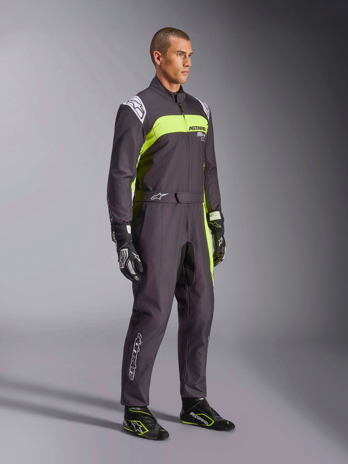 Męski model w kombinezonie Alpinestars KMX-9 V3 Graphic 3 w kolorze czarnym i żółtym fluorescencyjnym, profesjonalny strój kartingowy z jaskrawymi neonowymi żółtymi panelami i białymi akcentami logo, z dopasowanymi rękawicami i butami wyścigowymi, na tle studyjnym.