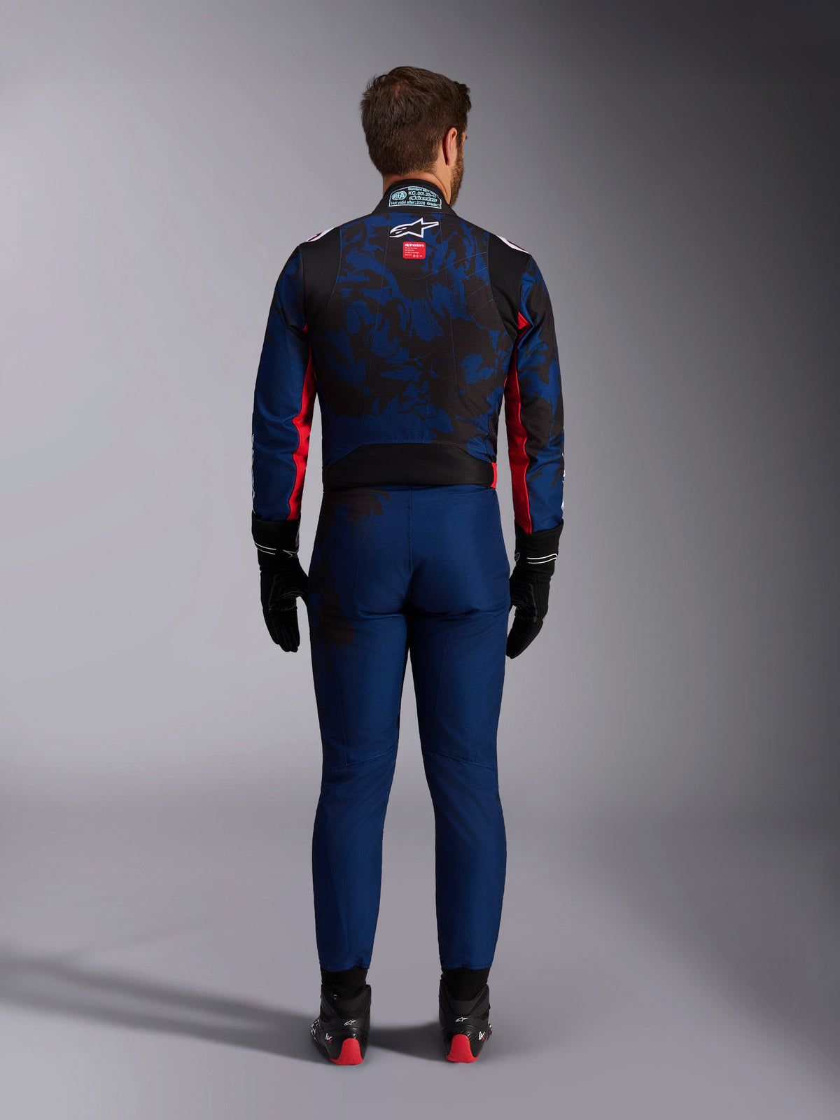 Kierowca kartingowy widoczny od tyłu w kombinezonie KMX-9 V3 Graphic 5 w kolorze Navy Red, granatowo-czarny wzór graficzny, czerwone boczne akcenty i logo Alpinestars na plecach, czarne rękawice i buty wyścigowe