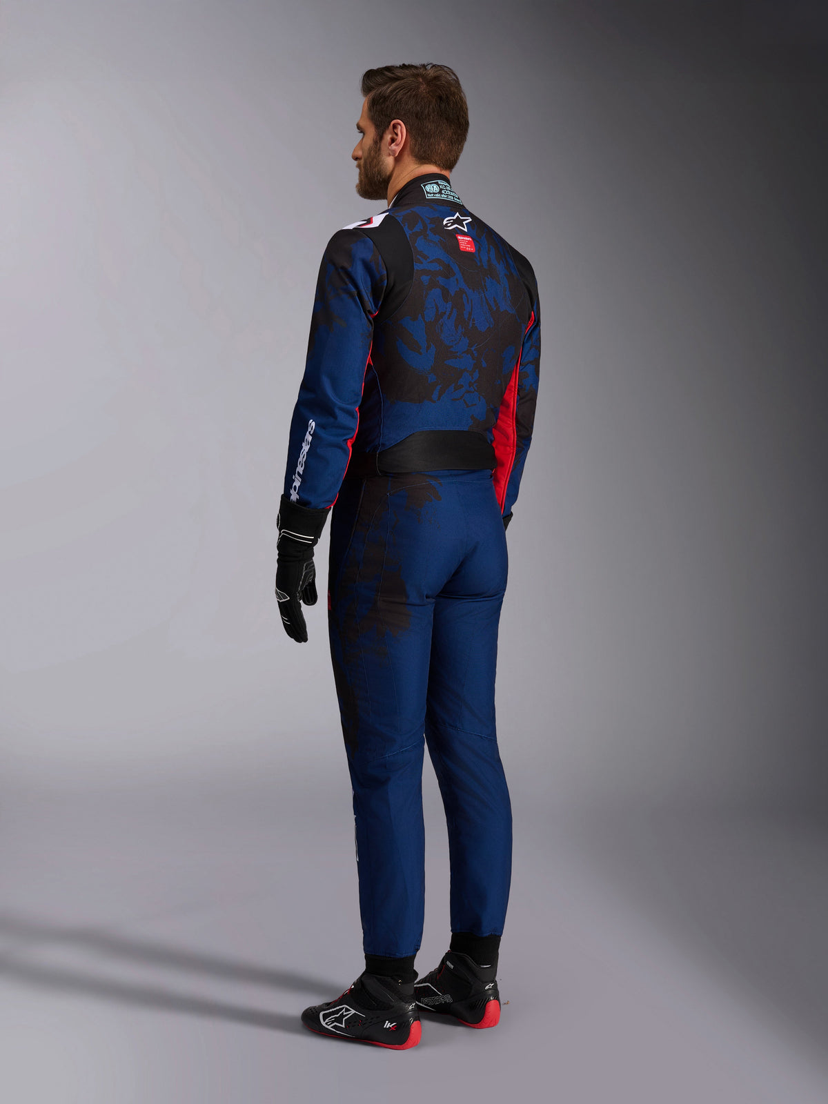 Alpinestars KMX-9 V3 Graphic 5 Suit, kombinezon kartingowy, granatowo-czerwony, widok z tyłu pokazujący granatowy korpus z ciemną grafiką inspirowaną kamuflażem na plecach i czerwonymi akcentami, noszony z czarnymi rękawicami wyścigowymi i czarno-czerwonymi butami kartingowymi