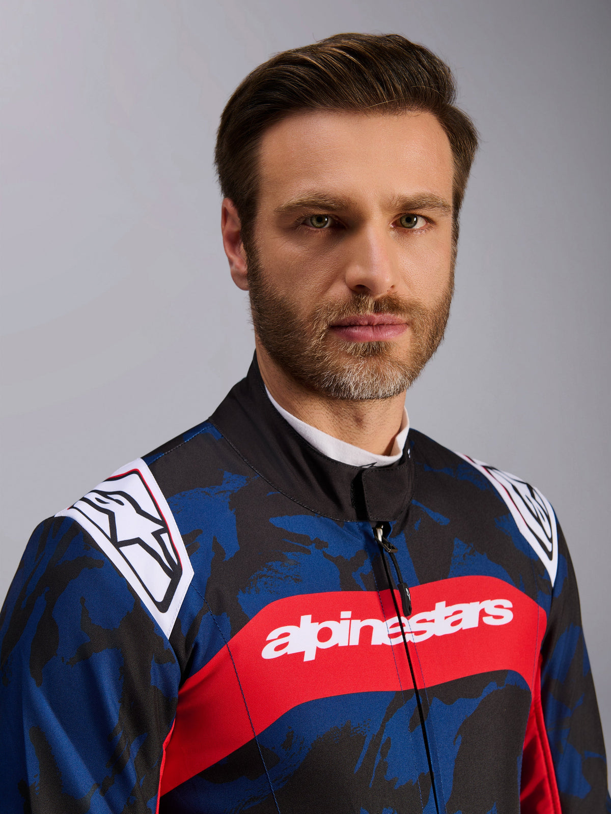 Kombinezon Alpinestars KMX-9 V3 Graphic 5, kombinezon kartingowy w kolorze granatowo-czerwonym, z niebiesko-czarnym wzorem kamuflażu, wyrazisty czerwony poziomy pas z białym napisem alpinestars na klatce piersiowej oraz białe logo na ramionach, prezentowany przez modela w studiu
