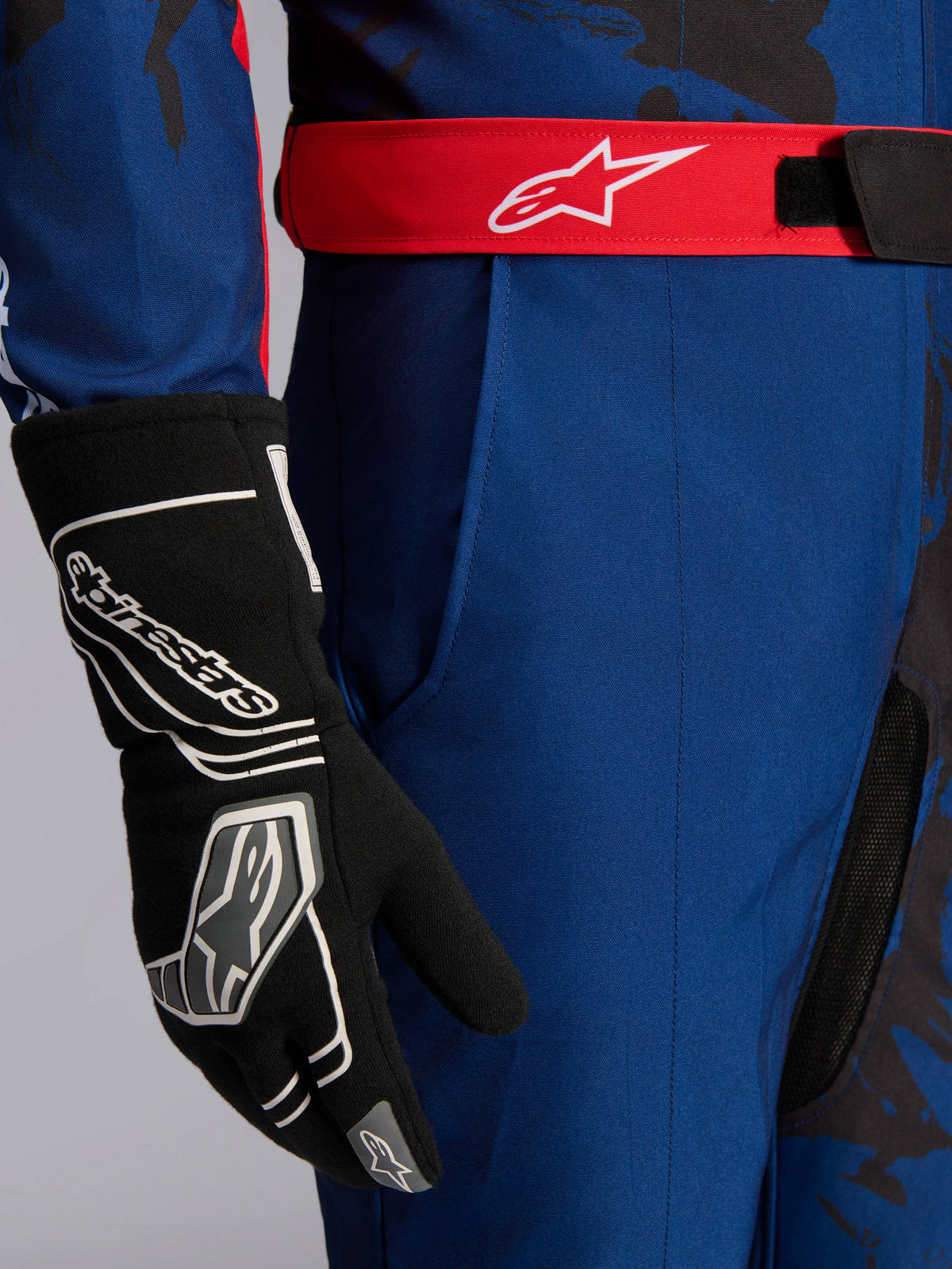 Alpinestars KMX-9 V3 Graphic 5 Suit, kombinezon kartingowy, granatowy i czerwony, zbliżenie na czerwony regulowany pas z białym logo, czarna rękawica kartingowa, wytrzymała granatowa tkanina i zintegrowana kieszeń na udzie