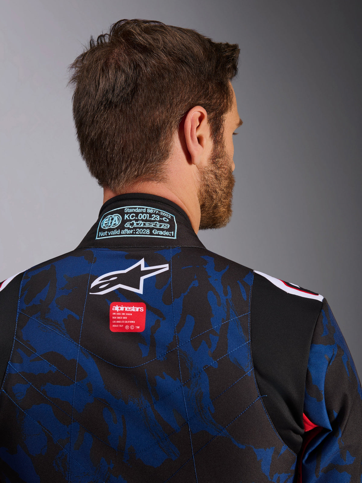 Zbliżenie na kombinezon kartingowy Alpinestars KMX-9 V3 Graphic 5 w kolorze granatowo-czerwonym, z niebiesko-czarnym wzorem kamuflażu, białym logo gwiazdy i czerwoną metką marki w górnej części pleców oraz naszywką certyfikacji FIA 8877-2022 na kołnierzu.