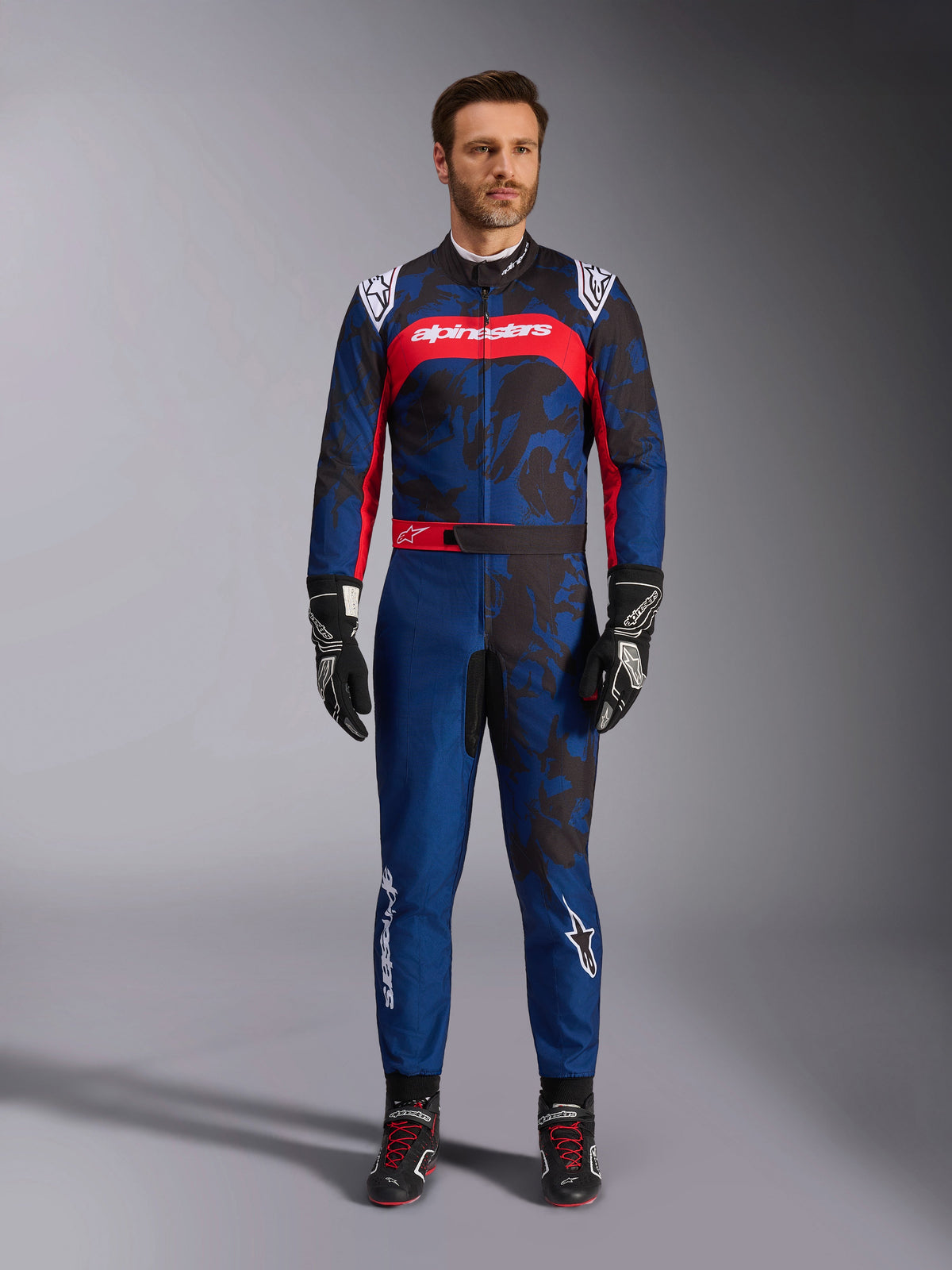 Alpinestars KMX-9 V3 Graphic 5 Suit, kombinezon kartingowy w kolorze granatowym i czerwonym, z abstrakcyjnym wzorem graficznym w stylu kamuflażu, prezentowany przez modela w czarnych rękawicach i butach wyścigowych, sportowy krój z wyraźnym logo Alpinestars na klatce piersiowej i nogawkach