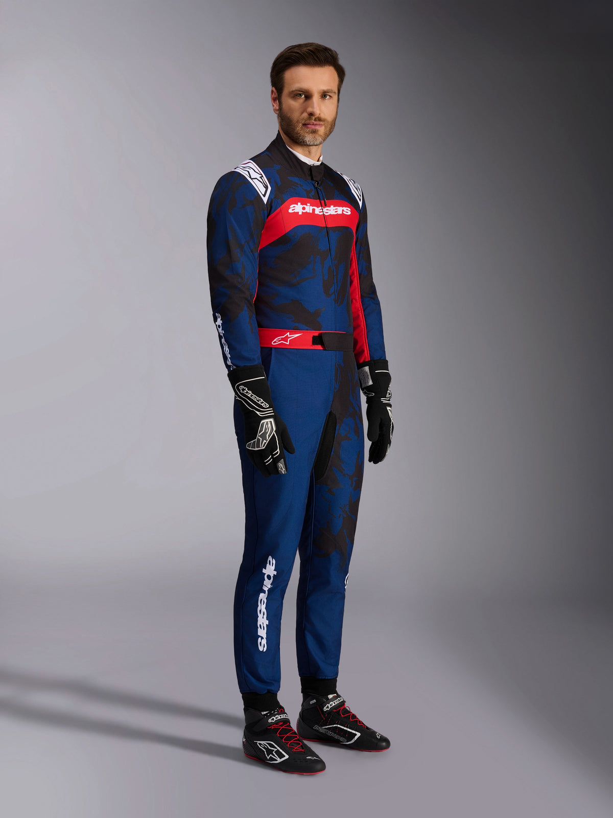 Model w kombinezonie Alpinestars KMX-9 V3 Graphic 5 w kolorze Navy Red, ujęcie całej sylwetki, czarne rękawice kartingowe i czarne buty wyścigowe, granatowy kombinezon z ciemną grafiką w stylu kamuflażu, czerwony panel na klatce piersiowej z białym logo, profesjonalny kartingowy strój ochronny