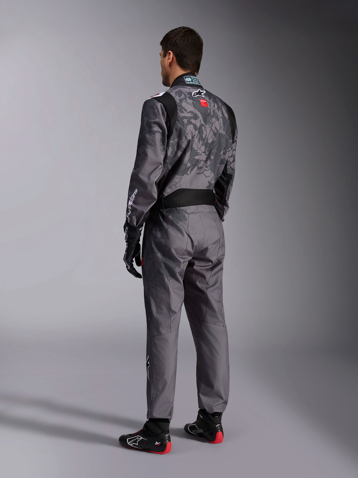 Alpinestars KMX-9 V3 Graphic 5 Suit, ciemnoszary i czarny kombinezon kartingowy, widok z tyłu modelu w pełnym zestawie wyścigowym, w tym czarno-czerwone rękawice i buty kartingowe, ze zintegrowanymi wzorami graficznymi i brandingiem na plecach.