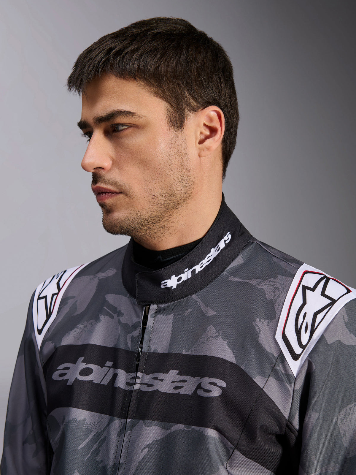 Alpinestars KMX-9 V3 Graphic 5 Suit, kombinezon kartingowy, wzór kamuflażu w kolorze ciemnoszarym i czarnym, zbliżenie na kołnierz i ramiona z białymi logo, profesjonalna odzież wyścigowa