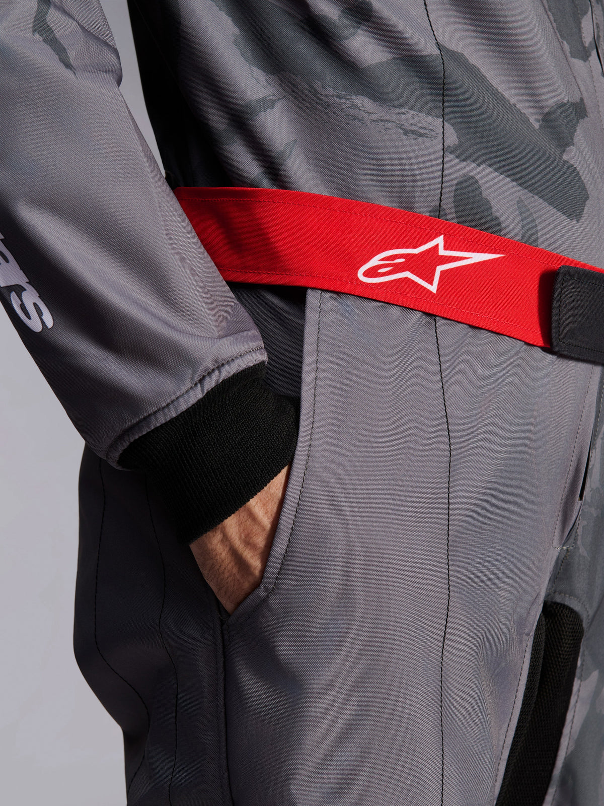 Alpinestars KMX-9 V3 Graphic 5 Suit, kombinezon kartingowy, ciemnoszary i czarny z czerwonym pasem, zbliżenie na czerwony regulowany pas w talii z białym logo, dłoń w kieszeni na udzie, czarny dzianinowy mankiet na szarym materiale z graficznym wzorem