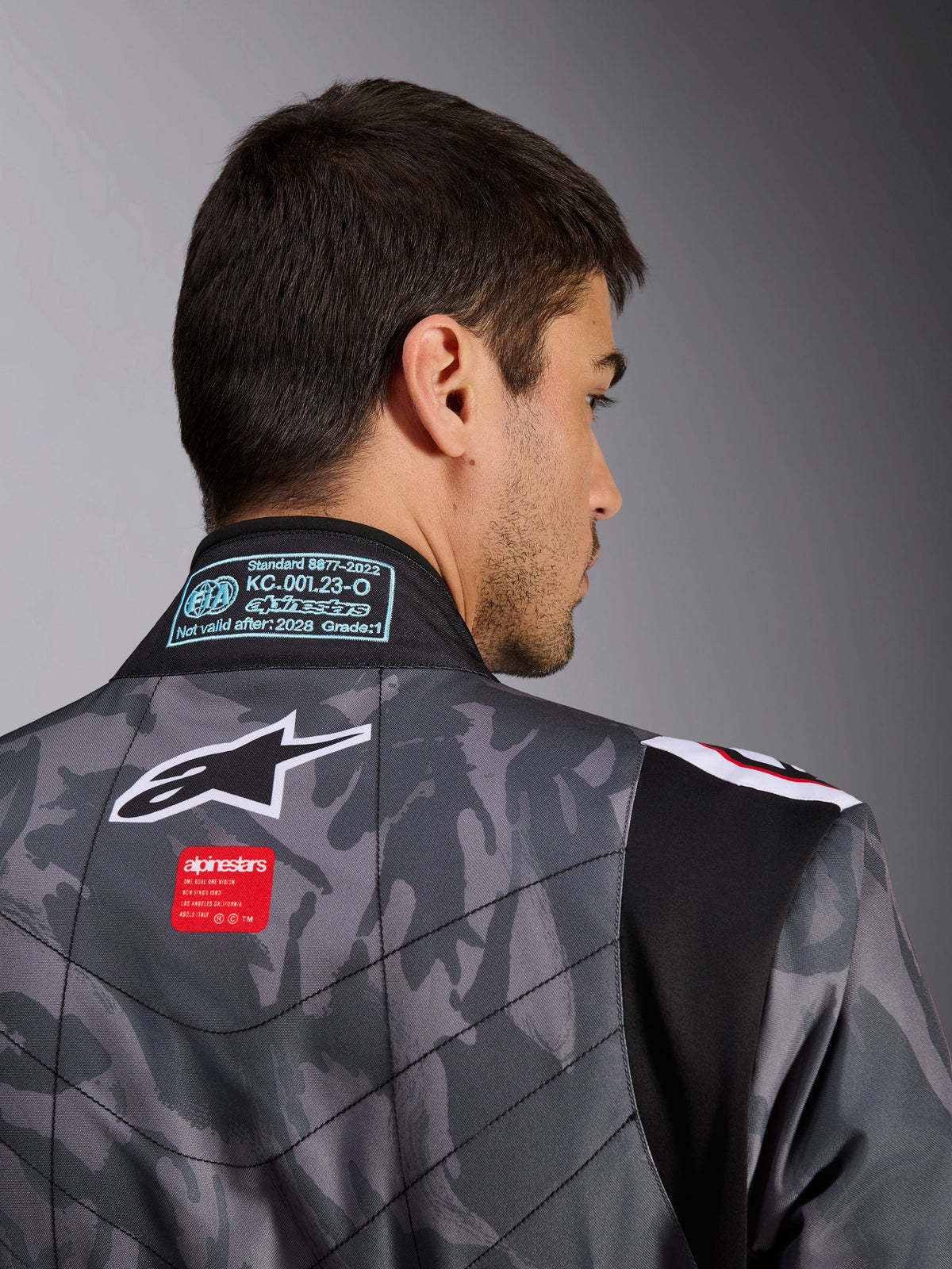 Alpinestars KMX-9 V3 Graphic 5 Suit, kombinezon kartingowy, wzór kamuflażu w kolorze ciemnoszarym i czarnym, zbliżenie na górną część pleców pokazujące logo Alpinestars, czerwoną metkę brandingową i etykietę homologacji FIA na kołnierzu.