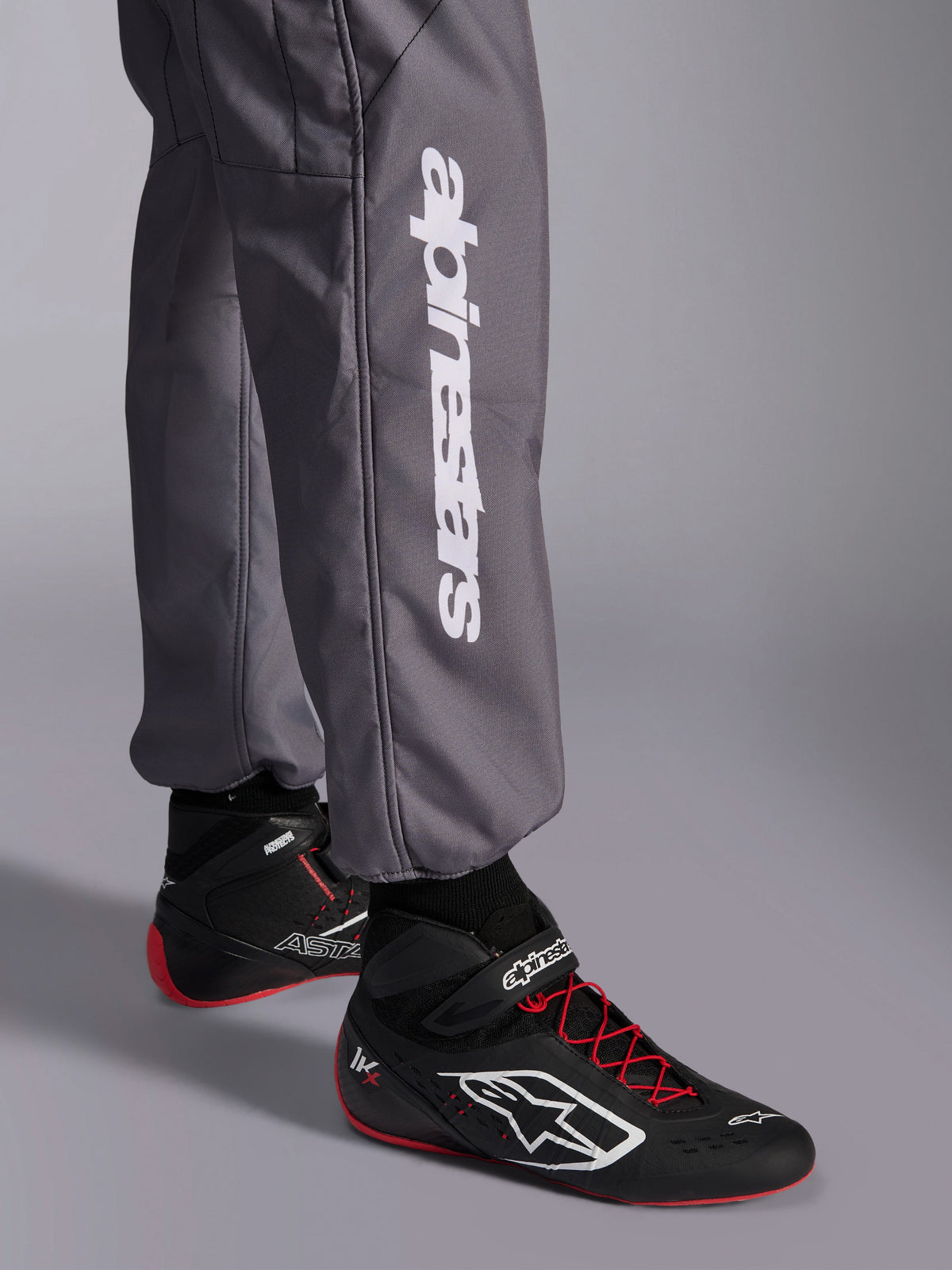 Alpinestars KMX-9 V3 Graphic 5 Suit, kombinezon kartingowy, ciemnoszary, zbliżenie na dolną część nogawki z białym pionowym logo Alpinestars, noszony z czarno-czerwonymi butami wyścigowymi, wytrzymały materiał do zawodów kartingowych