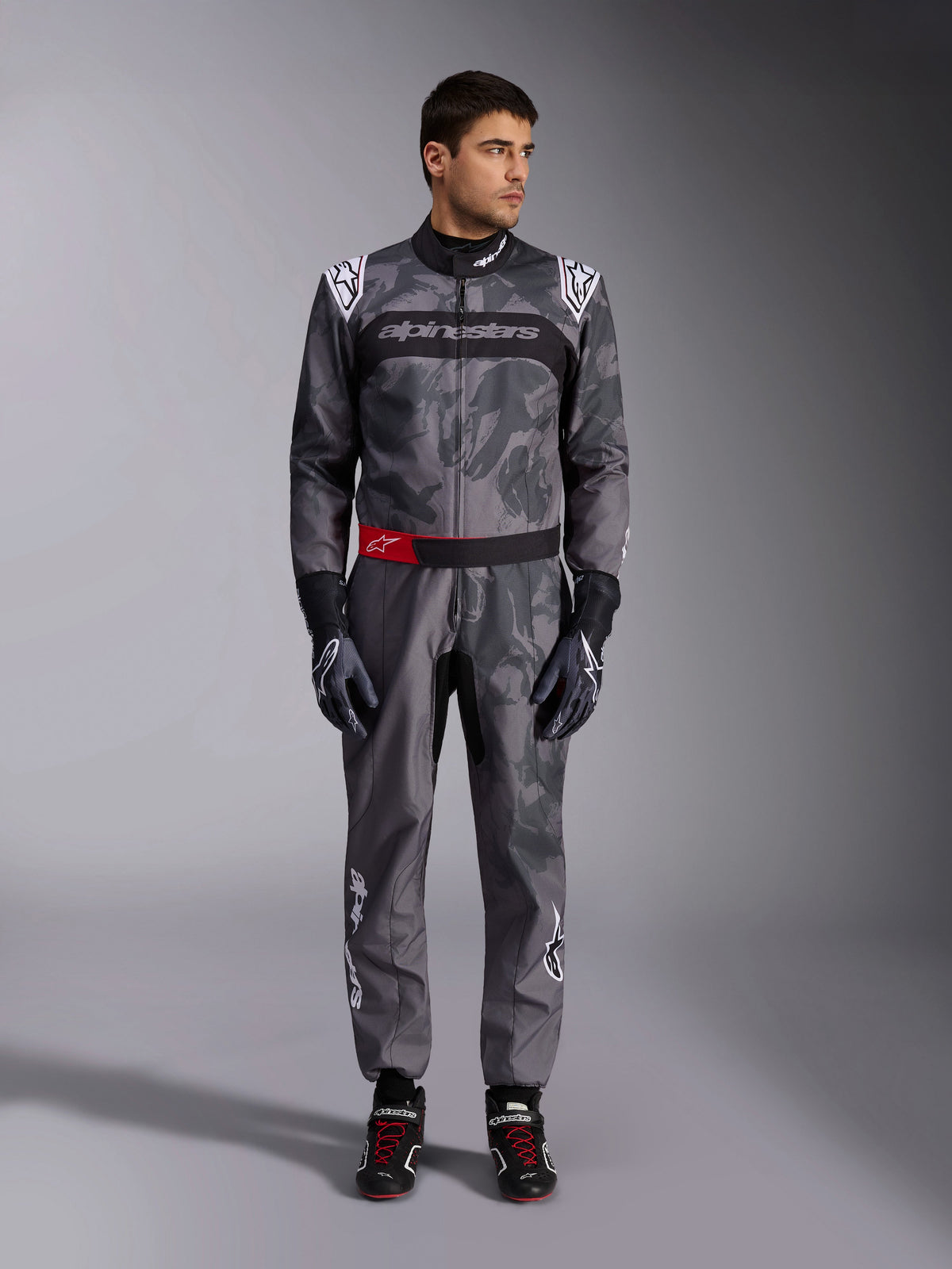 Alpinestars KMX-9 V3 Graphic 5 Suit, kombinezon kartingowy, ciemnoszary i czarny z grafikami w stylu kamuflażu, z czarnym pasem na klatce piersiowej z białym logo i czerwonym akcentem paska w talii, noszony z dopasowanymi rękawicami i butami wyścigowymi, profesjonalna odzież motorsportowa