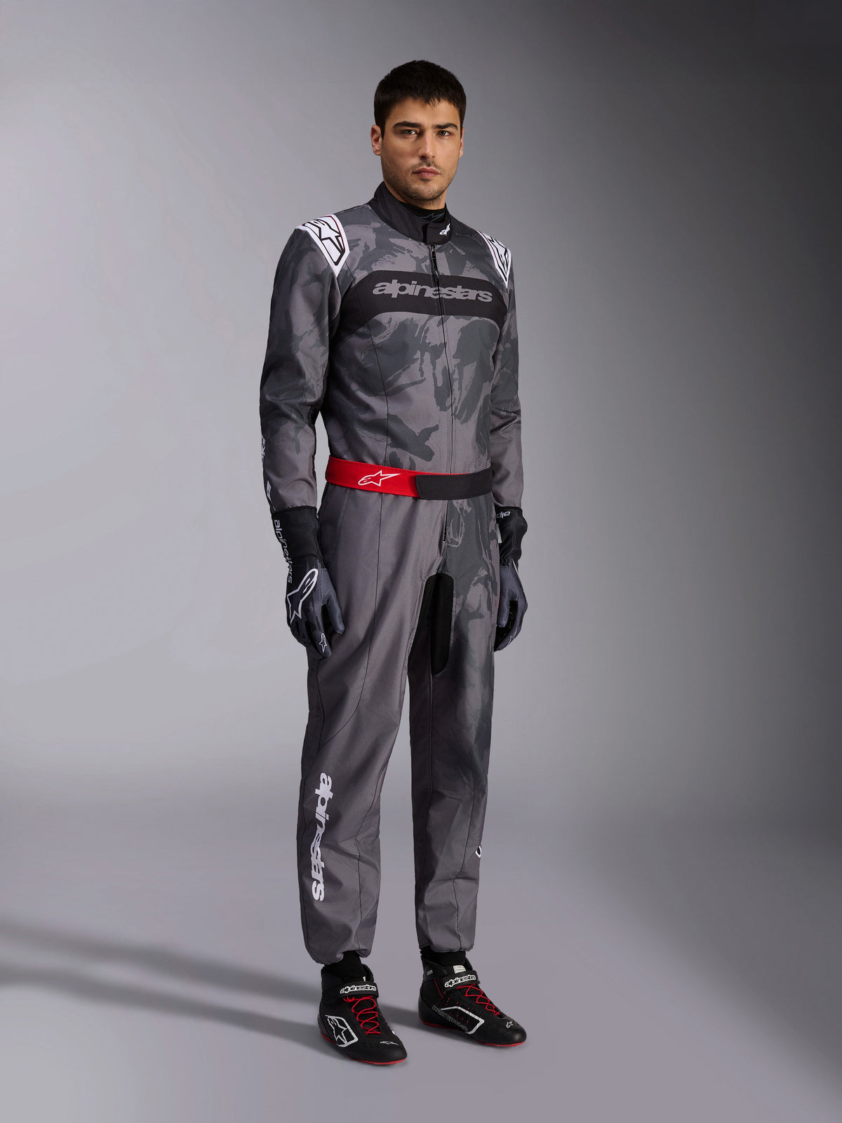 Kombinezon Alpinestars KMX-9 V3 Graphic 5, kombinezon kartingowy, ciemnoszary i czarny z graficznym wzorem kamuflażu, czerwony pas w talii z białym logo, noszony z rękawicami i butami wyścigowymi, profesjonalny sprzęt motorsportowy do kartingu