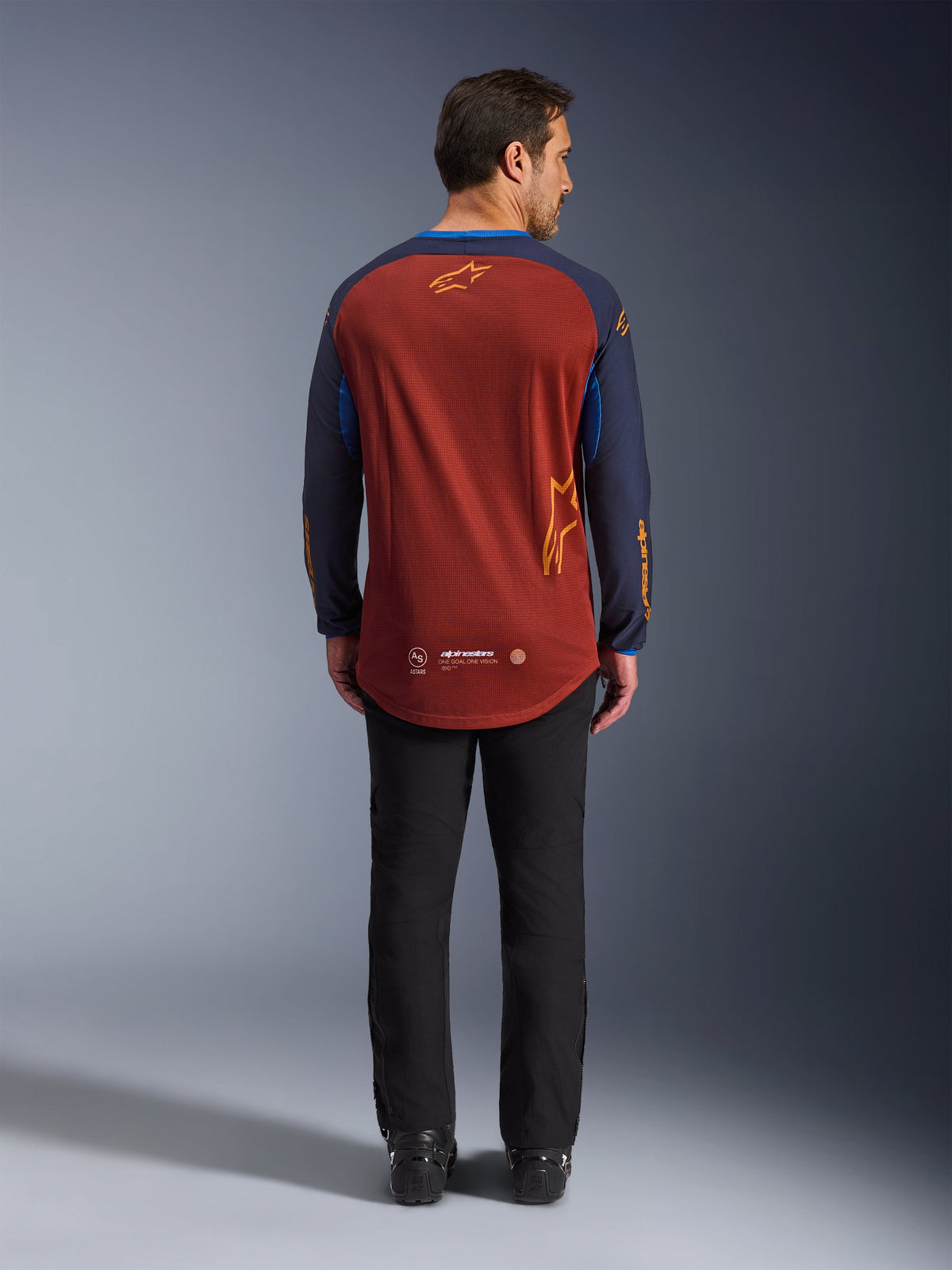 Alpinestars Maxdura Dual Jersey w kolorach Rust Brown i Mood Indigo, widok z tyłu pokazujący przedłużony tył i detale z logo, noszona z czarnymi spodniami technicznymi i butami, zaprojektowana do jazdy typu adventure i dual-sport