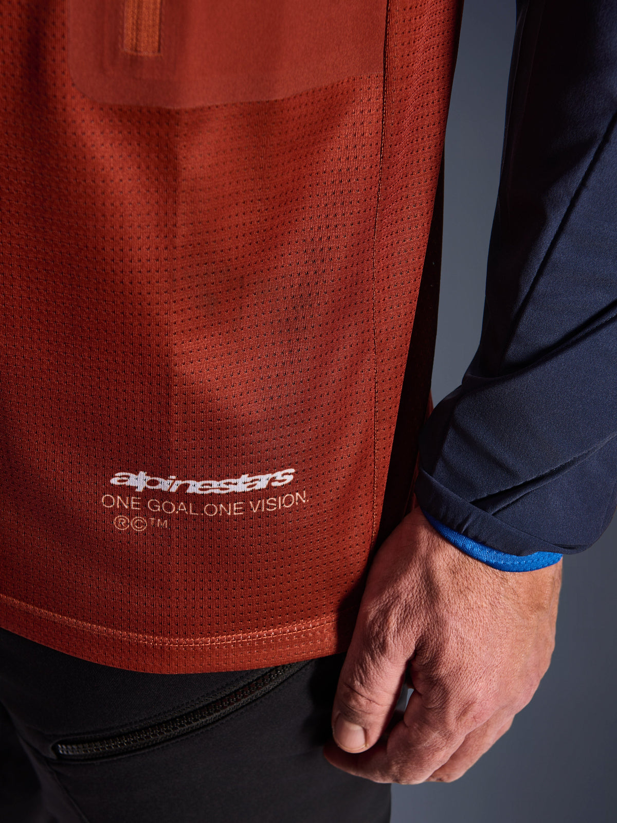 Alpinestars Maxdura Dual Jersey, koszulka motocyklowa Adventure, rdzawy brąz i granat Mood Indigo, zbliżenie na siatkową tkaninę tułowia i granatowy tkany rękaw, biały branding Alpinestars „One Goal One Vision” na dolnym brzegu