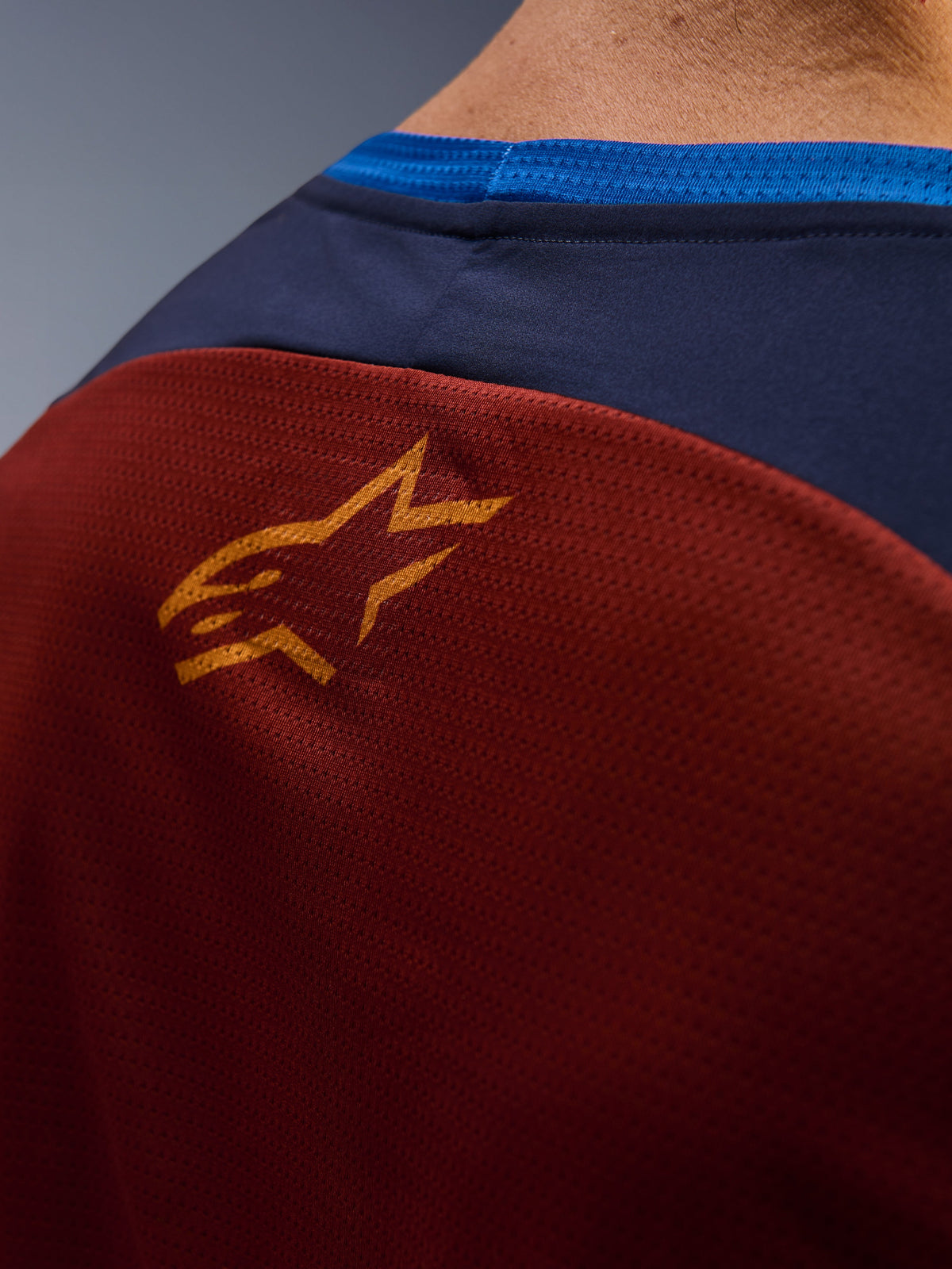 Alpinestars Maxdura Dual Jersey, zbliżenie na siatkowany materiał w kolorze Rust Brown na plecach, małe pomarańczowe logo gwiazdy, ciemnoniebieski górny panel i jasnoniebieskie wykończenie kołnierzyka, szczegółowy widok motocyklowej koszulki adventure