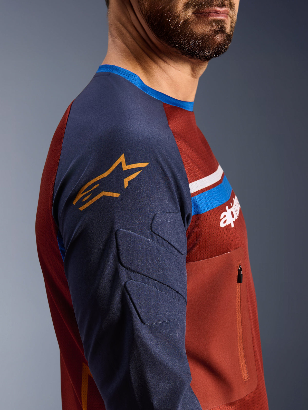 Koszulka Alpinestars Maxdura Dual, kolory Rust Brown i Mood Indigo, widok z profilu na wyściełane rękawy z żółtym logo gwiazdy, siatkowa konstrukcja tułowia z kieszenią na piersi na zamek, techniczna odzież motocyklowa do jazdy adventure