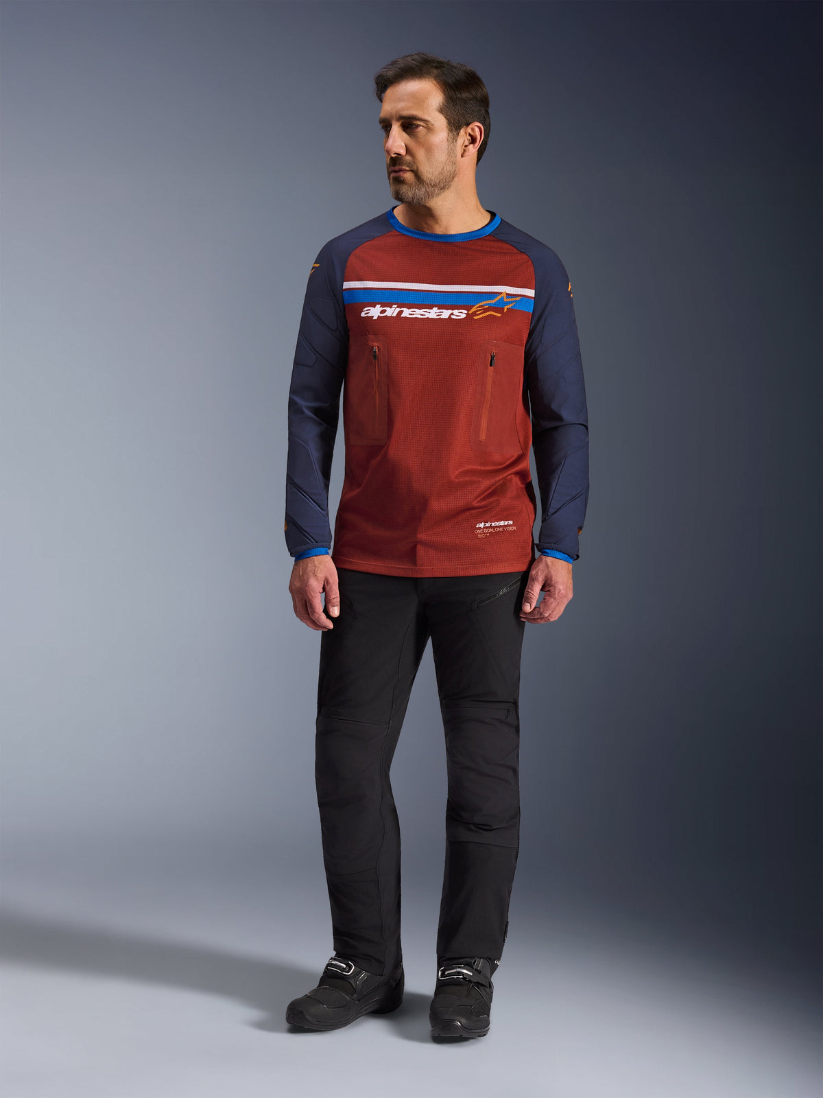 Alpinestars Maxdura Dual Jersey, rdzawy brąz i granat Mood Indigo, motocyklowa koszulka adventure z siatkowym tułowiem i granatowymi rękawami, model w czarnych spodniach i butach motocyklowych, techniczna odzież dual-sport do jazdy w terenie
